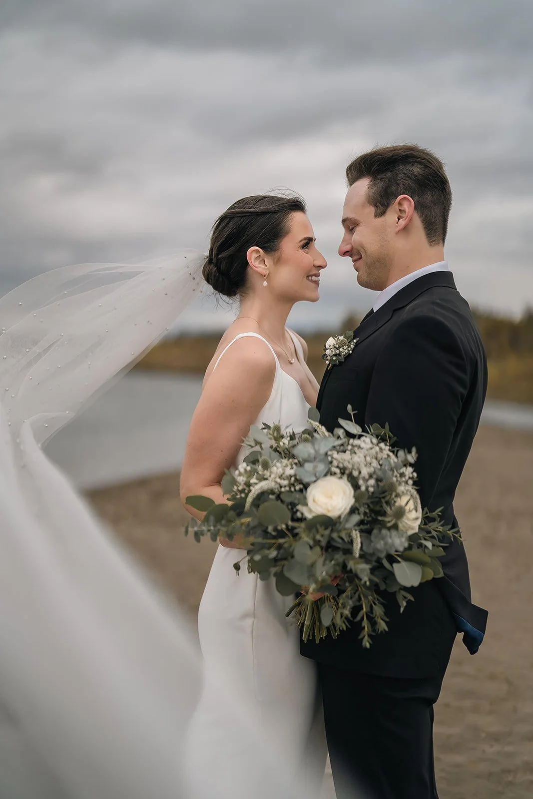 A bride and groom stand close together on a beach, smiling and looking at each other, with the bride holding a bouquet of white flowers and greenery, under a cloudy sky.