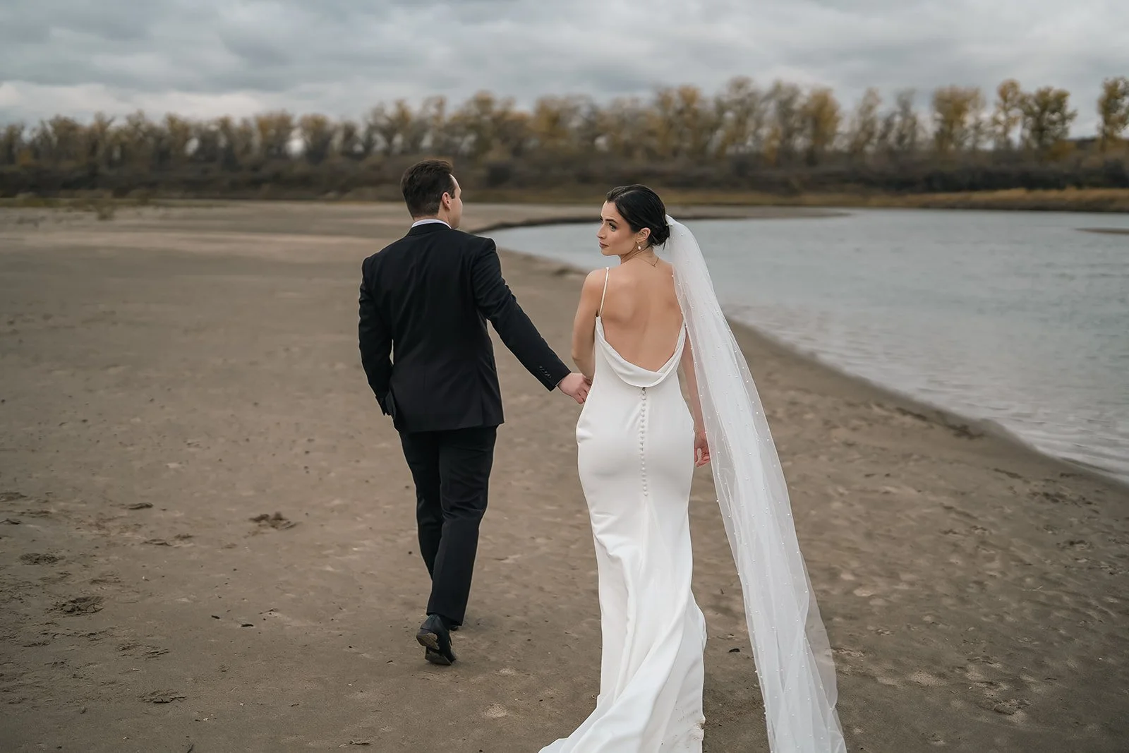 A bride and groom holding hands walking along a beach with water and trees in the background.