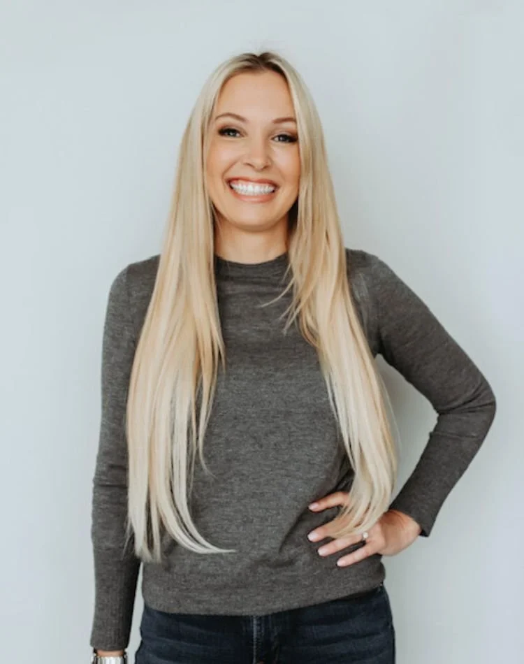 A smiling woman with long blonde hair, wearing a gray long-sleeve shirt and dark jeans, standing against a plain white background.