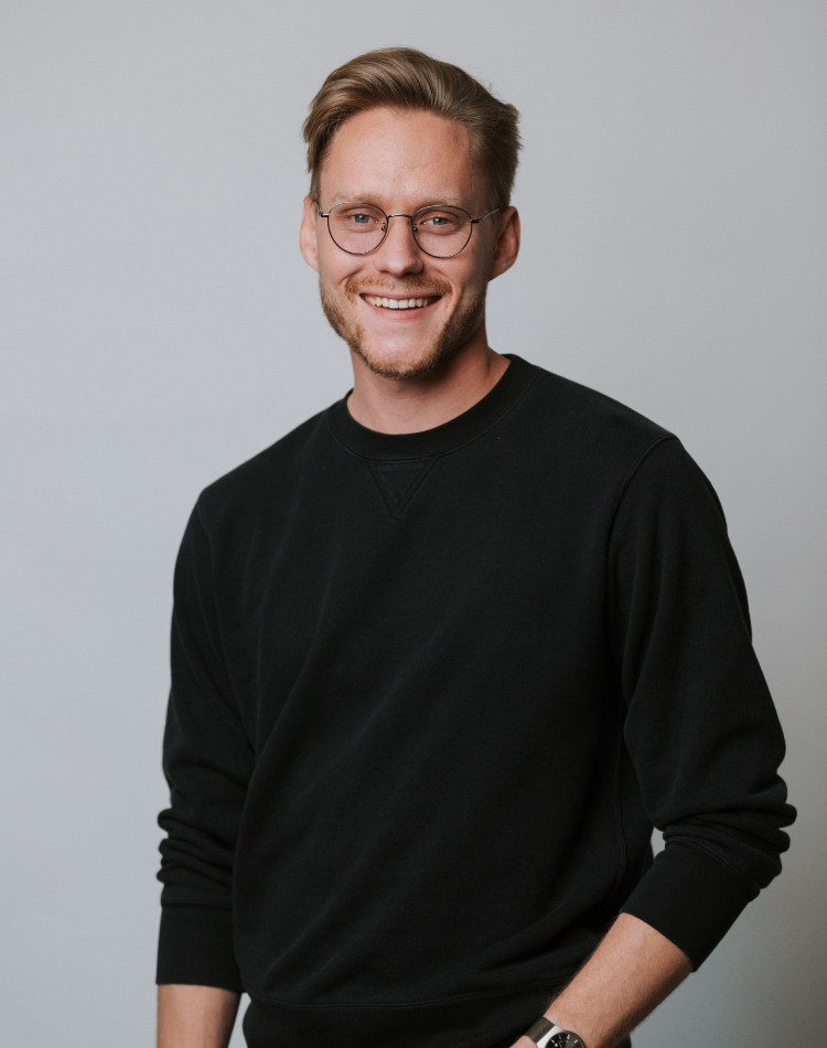 A young man with light brown hair, glasses, and a beard, smiling and wearing a black sweatshirt against a plain light gray background.