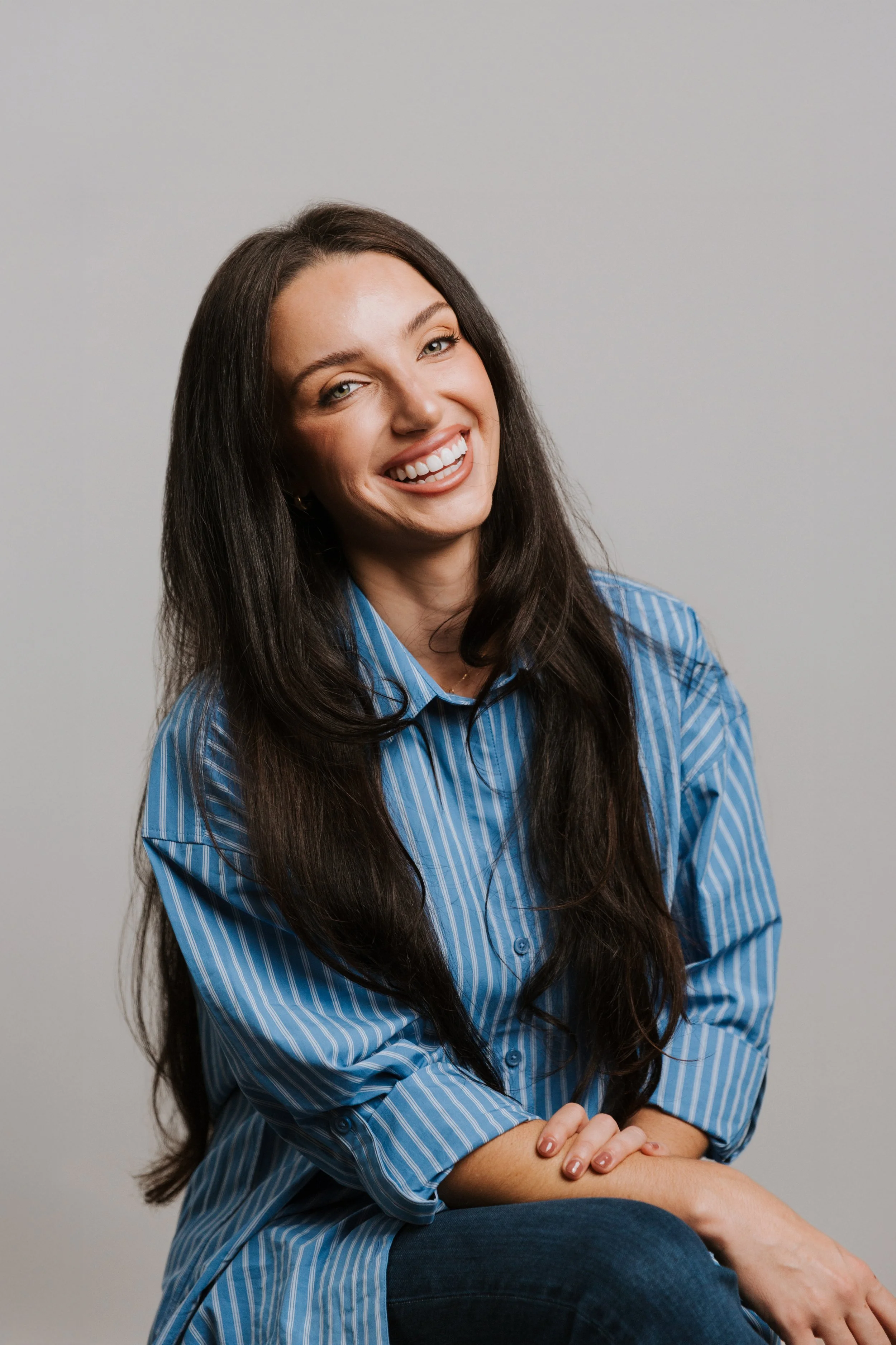 Rachel Burns, California virtual telehealth therapist specializing in teens and women. A woman with long dark hair, wearing a blue striped button-up shirt, smiling and sitting against a plain light gray background.