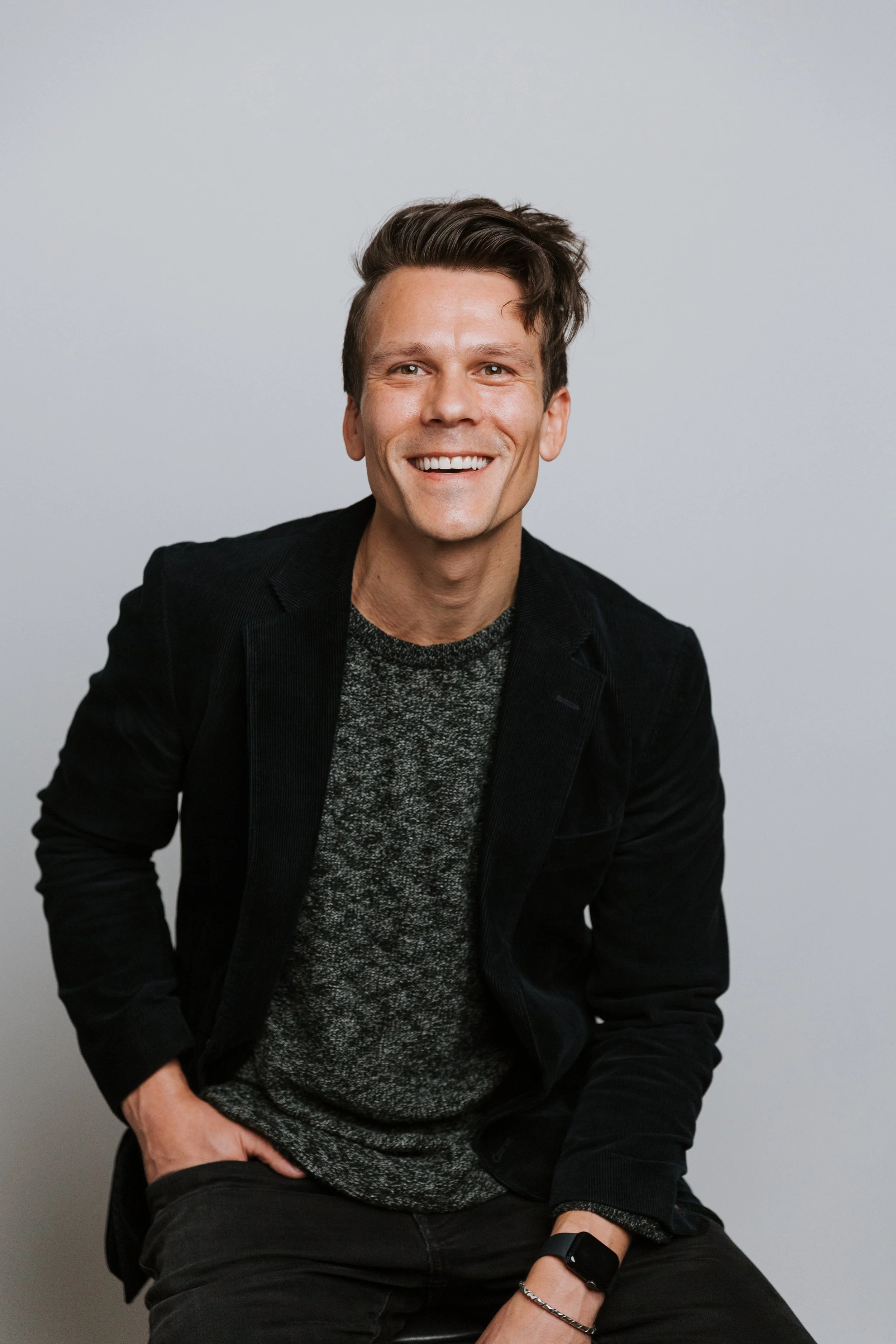 A smiling man with dark hair in a casual outfit, sitting against a plain light gray background.