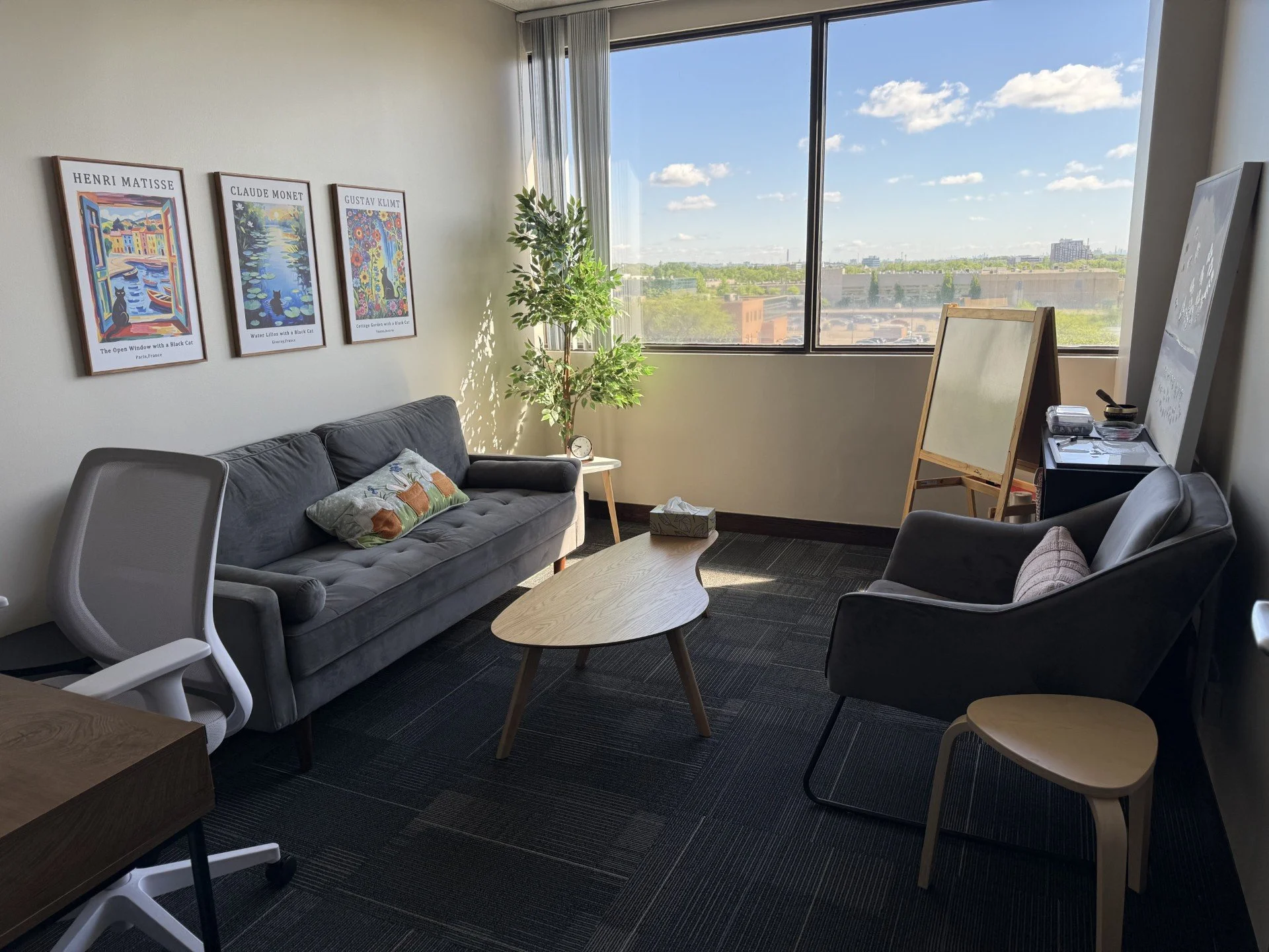 Office lounge with a gray sofa, a white chair, and a light wood coffee table. The wall has framed art posters of famous painters. A plant and a small clock are on a side table. Large windows show blue sky and cityscape outside.