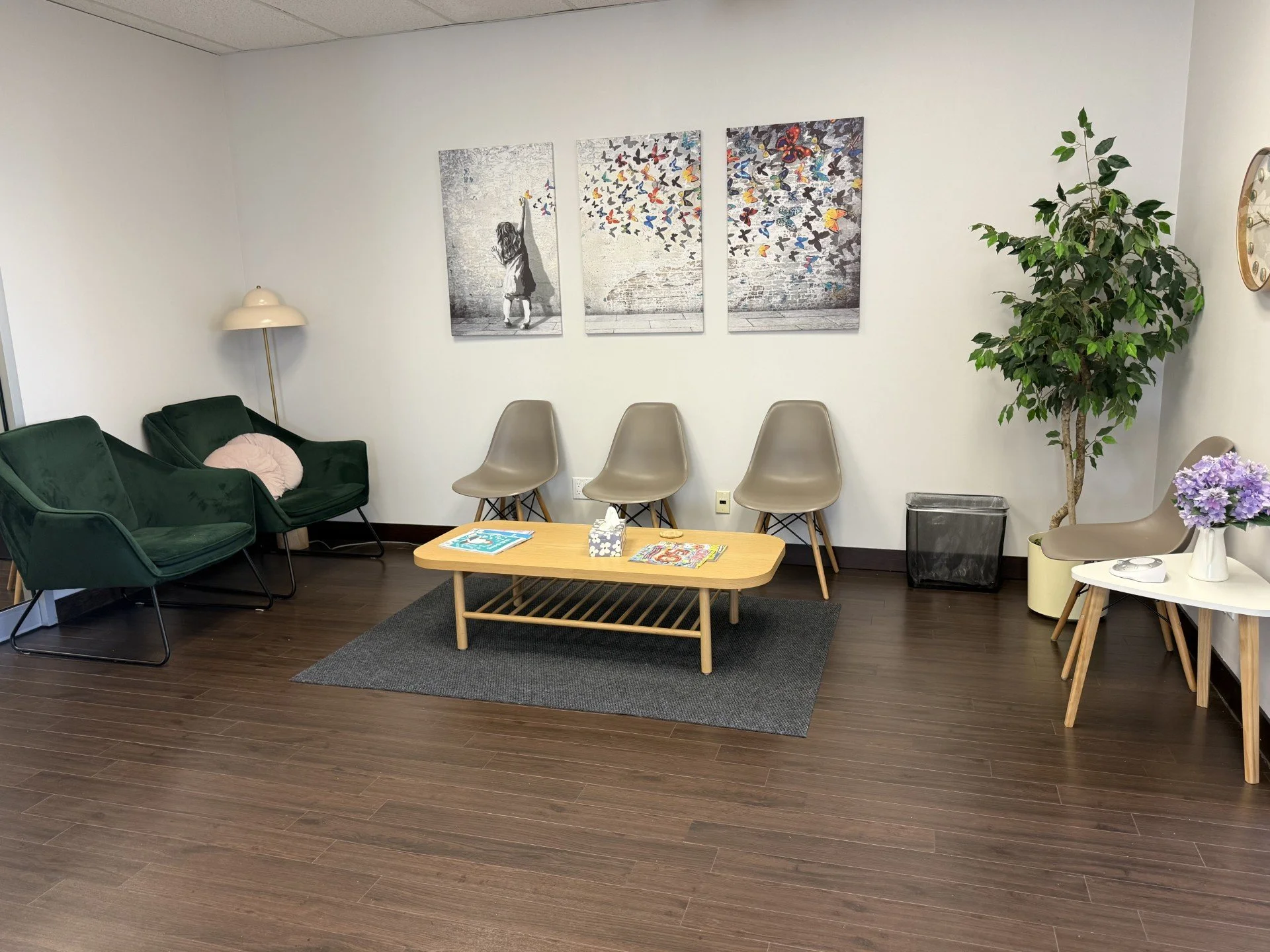 Waiting room with dark wood floor, green velvet sofa, beige chairs, a wooden coffee table holding magazines and tissue box, a gray rug, potted plant, wall art of a girl and colorful butterflies, white walls, clock, white side table with purple flowers, and a trash bin.