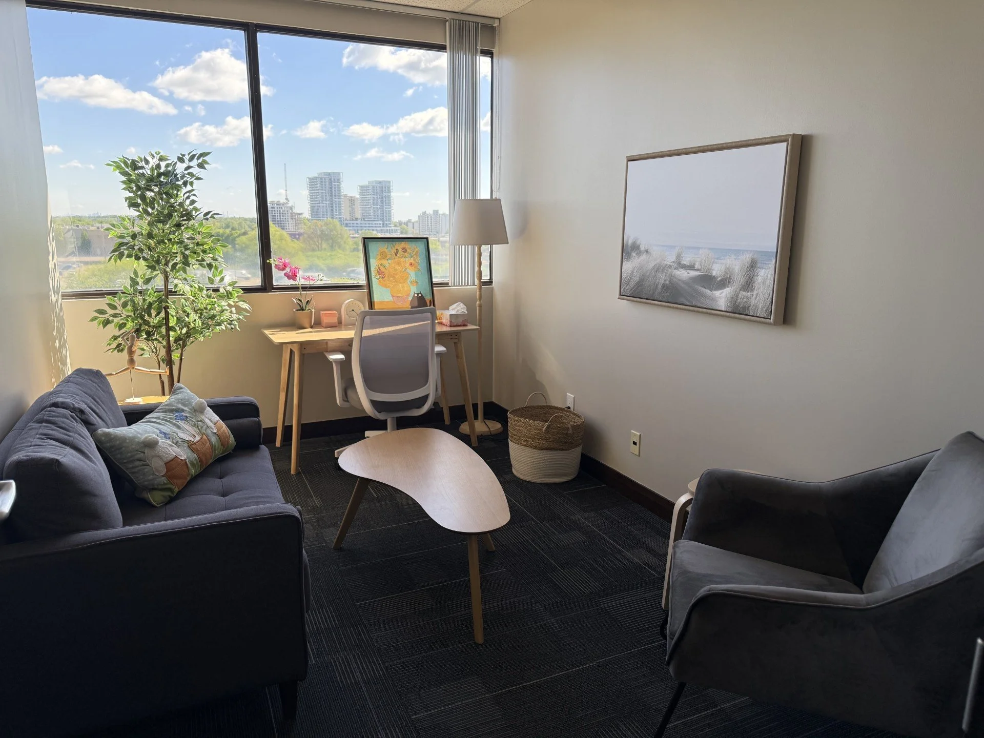 A cozy office with a large window showing a cityscape and blue sky with clouds. Inside, there's a small desk with a white office chair, a potted plant, a framed painting, a white lamp, and a tissue box. There are also two sofas and a coffee table, with wall art depicting a beach scene.