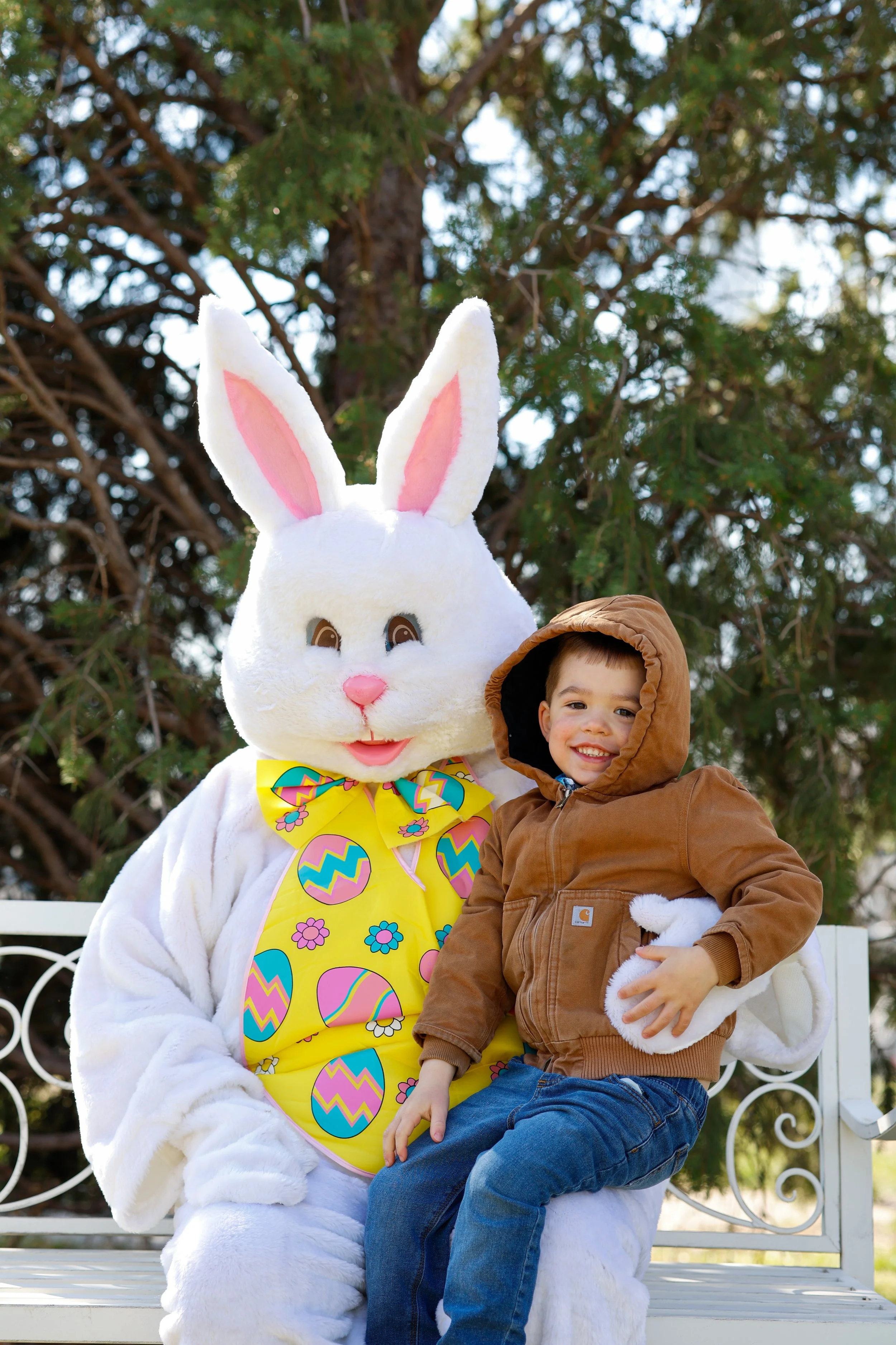 A young boy sitting on a white park bench next to a person in an Easter Bunny costume with a large head, pink nose, and colorful Easter egg apron. They are outdoors with a large evergreen tree in the background.