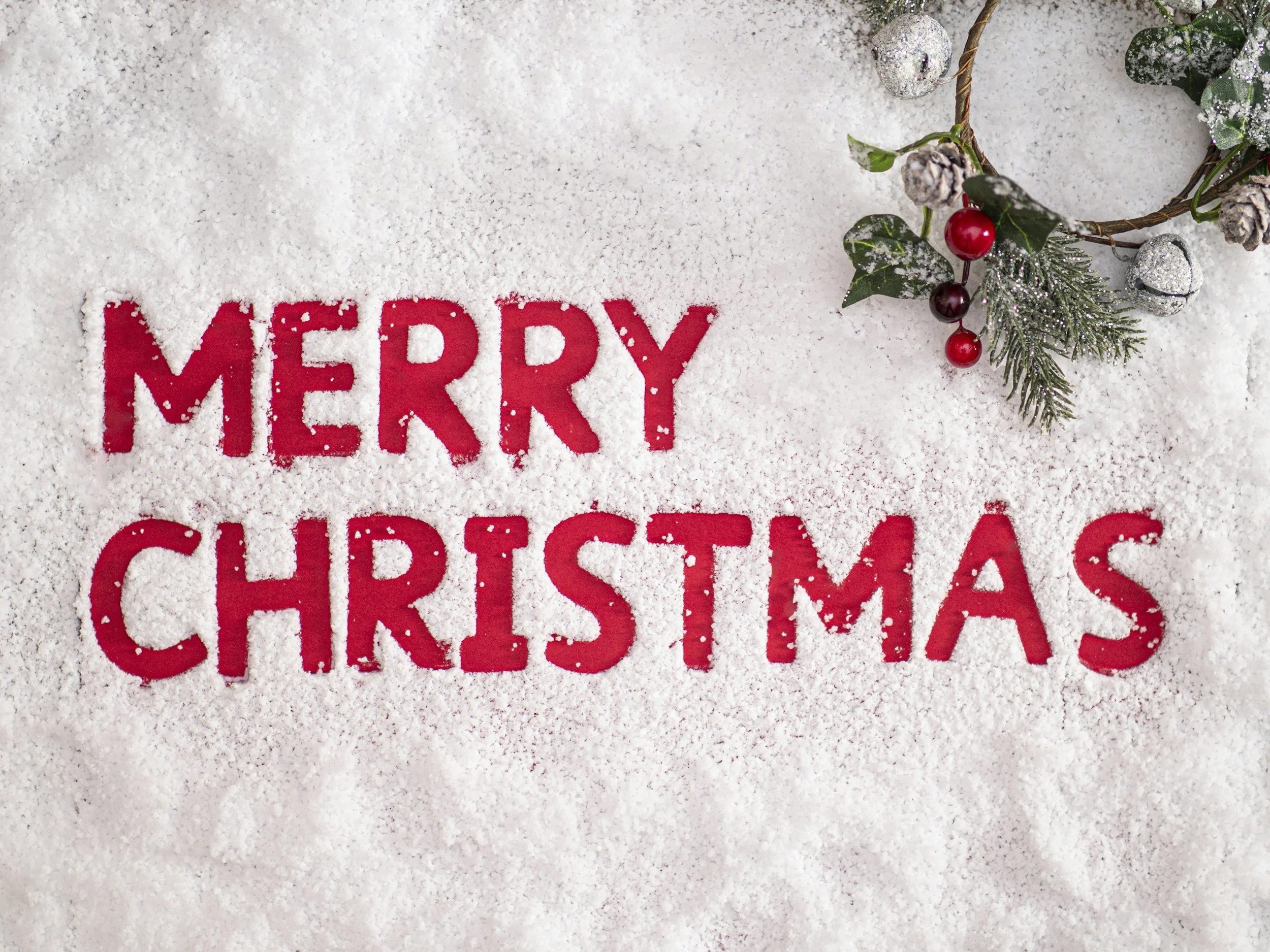 "Merry Christmas" text written in red on a snowy background with a festive wreath.