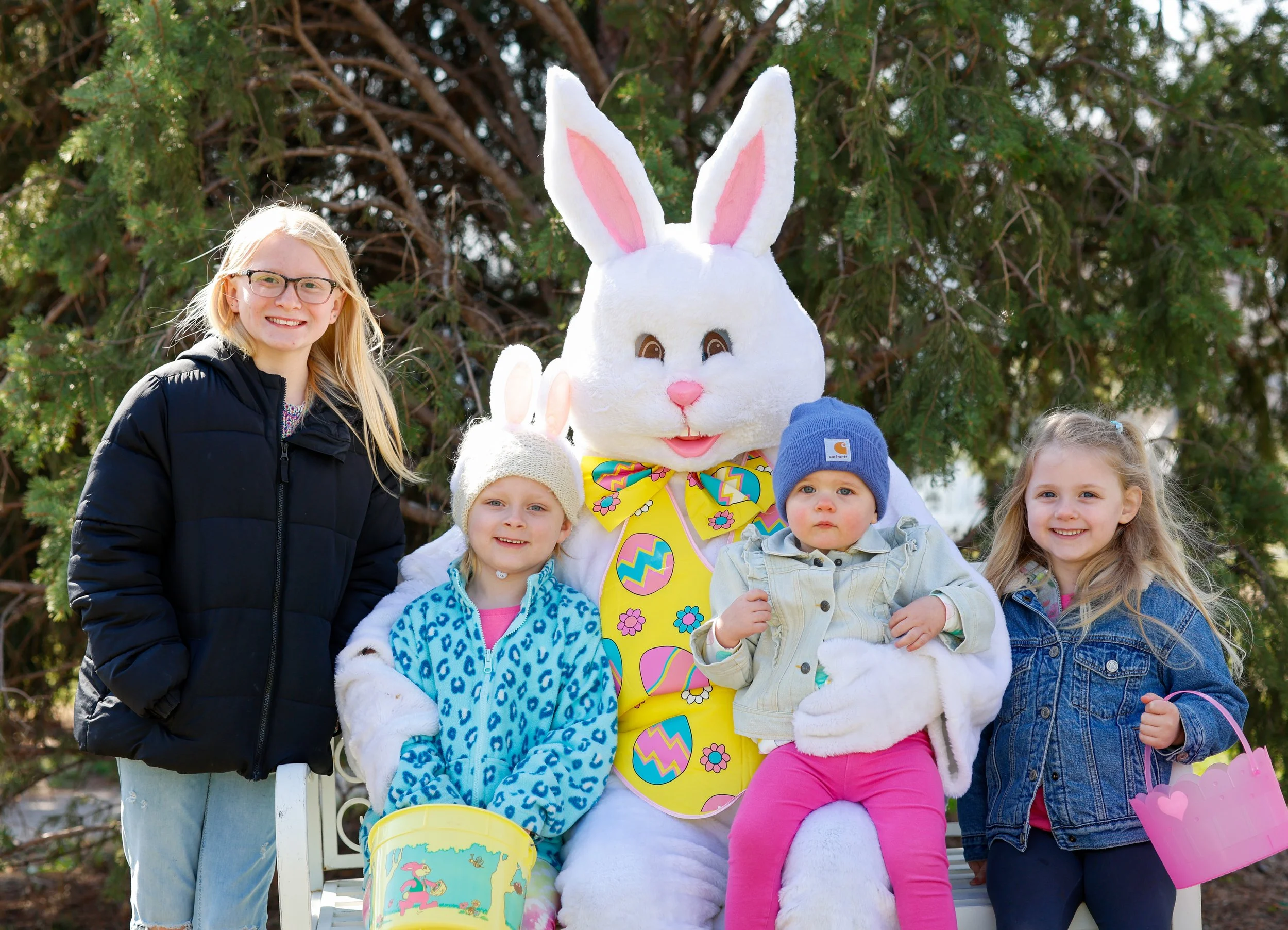 Children with a person in a large Easter bunny costume sitting on a park bench in front of a tree, celebrating Easter.