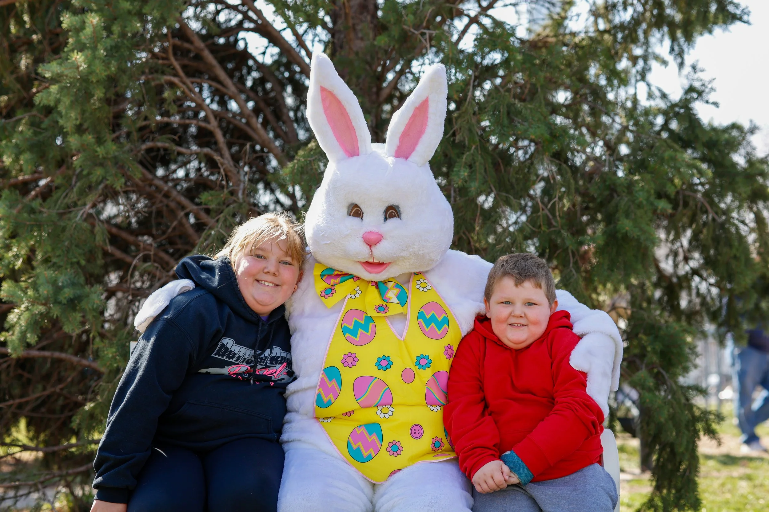 Two children sitting with an Easter Bunny mascot outdoors in front of a tree, smiling. The bunny costume is white with pink inner ears, a pink nose, and a yellow apron with colorful Easter eggs, flowers, and a bowtie. One child is wearing a blue hoodie, and the other is in a red hoodie.