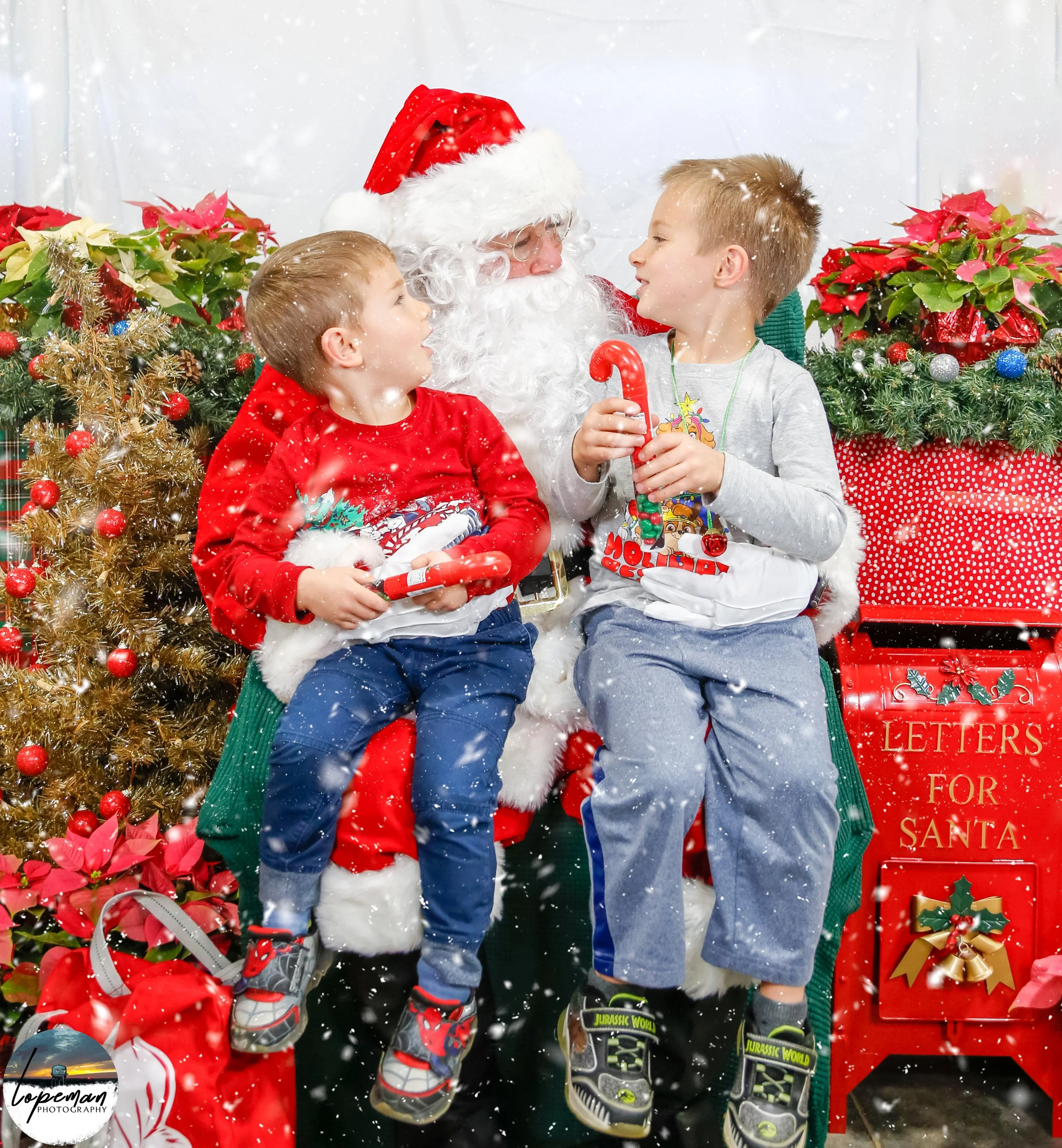 Two children sitting with Santa Claus surrounded by Christmas decorations, including poinsettias and a "Letters for Santa" mailbox, holding candy canes.