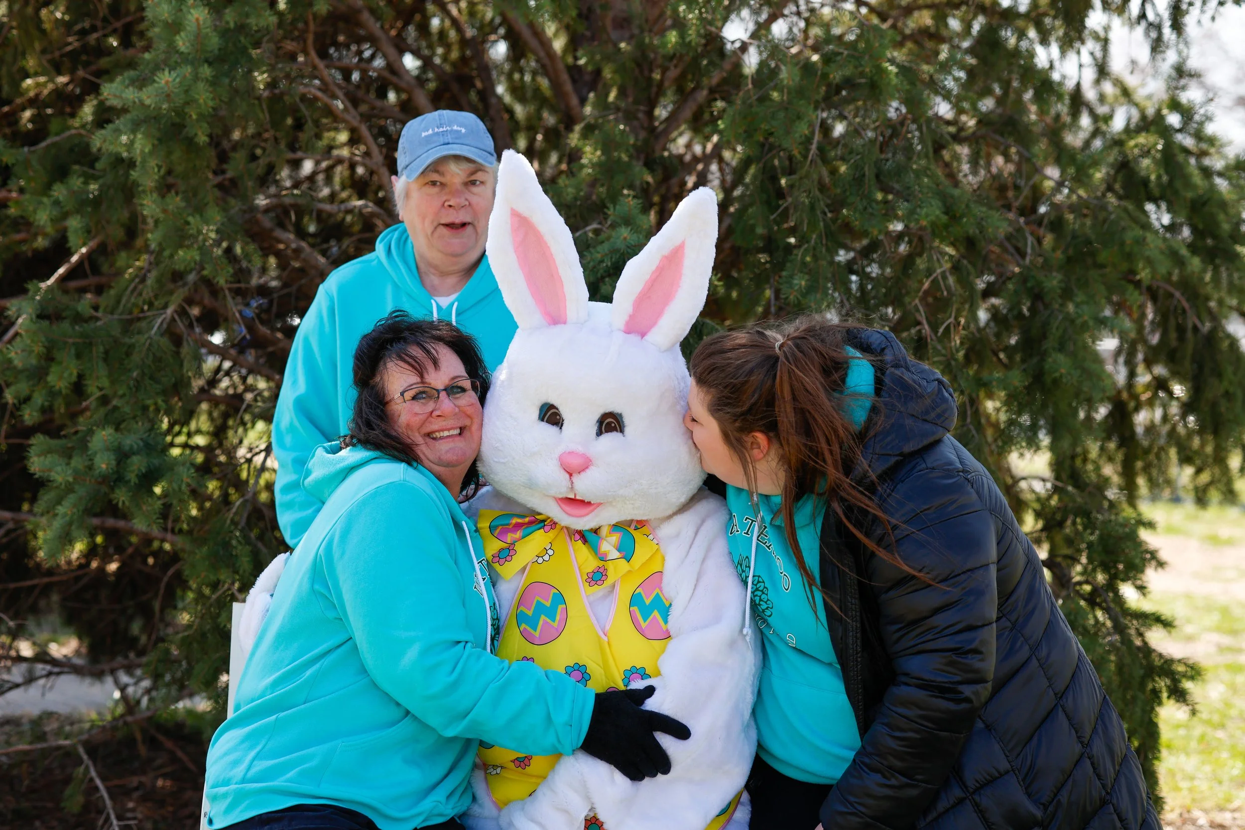 Three people and a person in a large Easter Bunny costume posing outdoors in front of a tree, with one person hugging the bunny and another leaning in for a kiss.