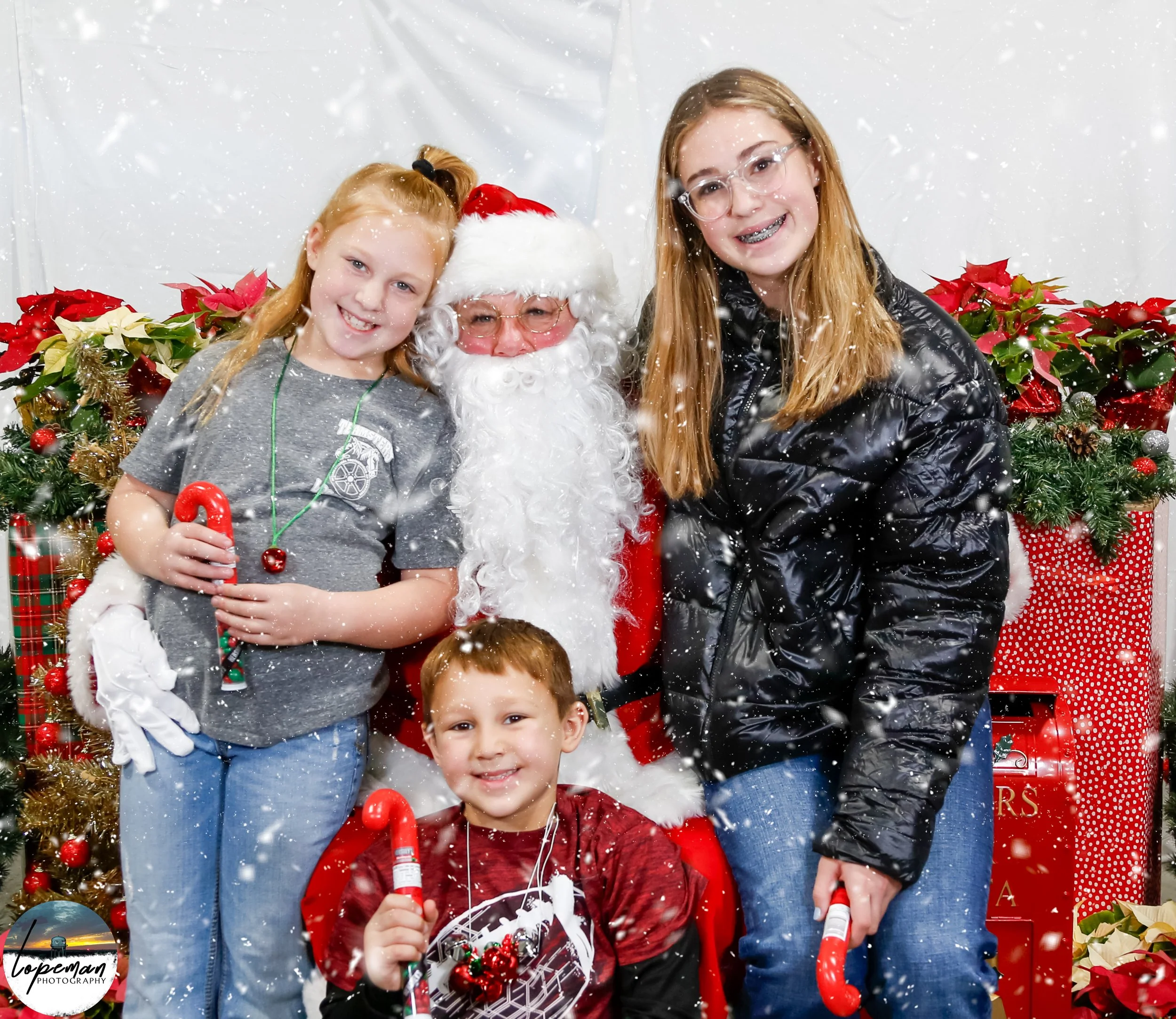 Santa Claus with three children holding candy canes, surrounded by holiday decorations and artificial snow.