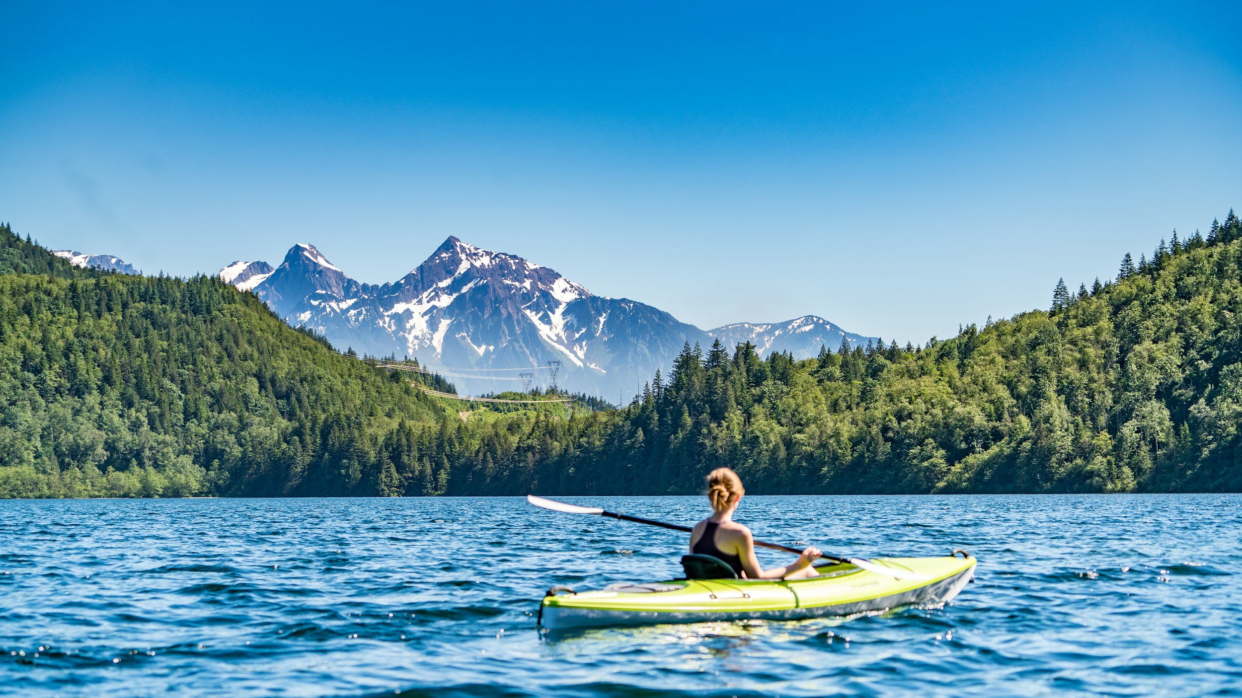 Person kayaking on a lake surrounded by lush green trees with snow-capped mountains in the background under a clear blue sky.