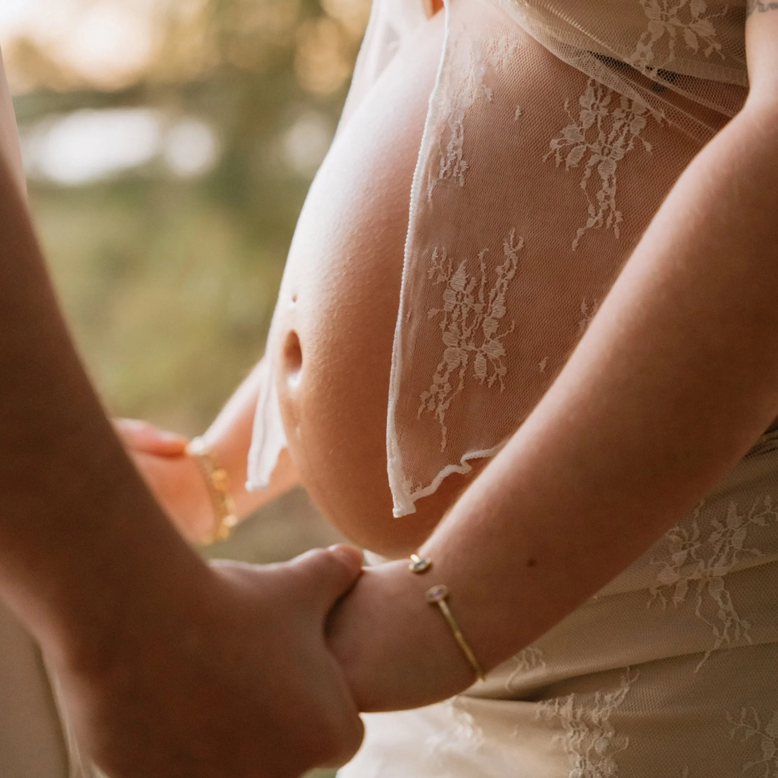 Close-up of a pregnant woman's belly, showing her belly button, with her hands gently cradling it. She is wearing a sheer, lace-trimmed garment, and there are blurred trees in the background.