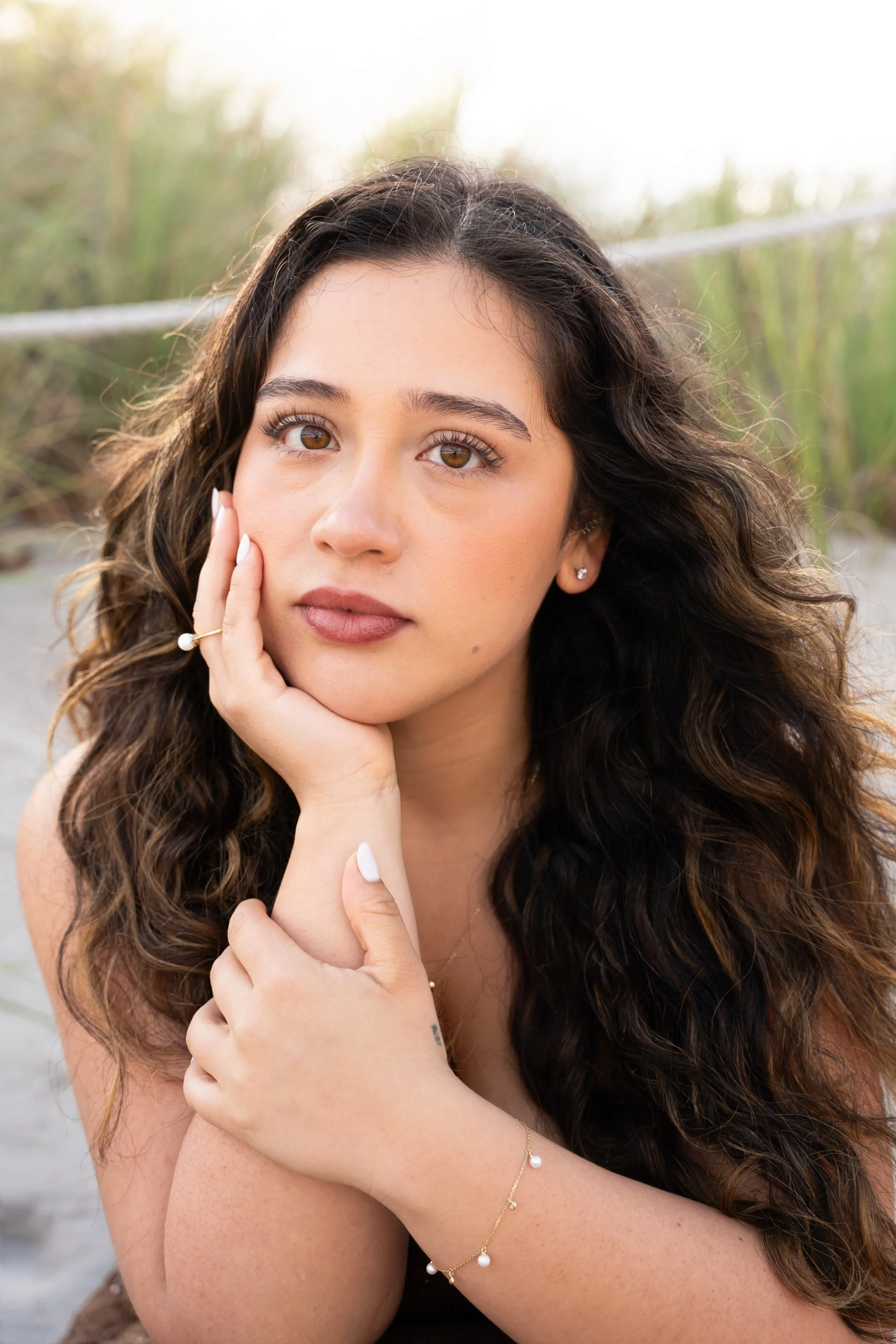 A young woman with long, curly brown hair, wearing small earrings and jewelry, resting her chin on her hand while posing outdoors in a natural setting with tall grass in the background.