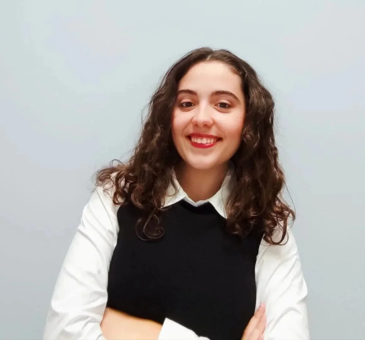 Young woman with curly brown hair smiling in front of a plain light gray background.