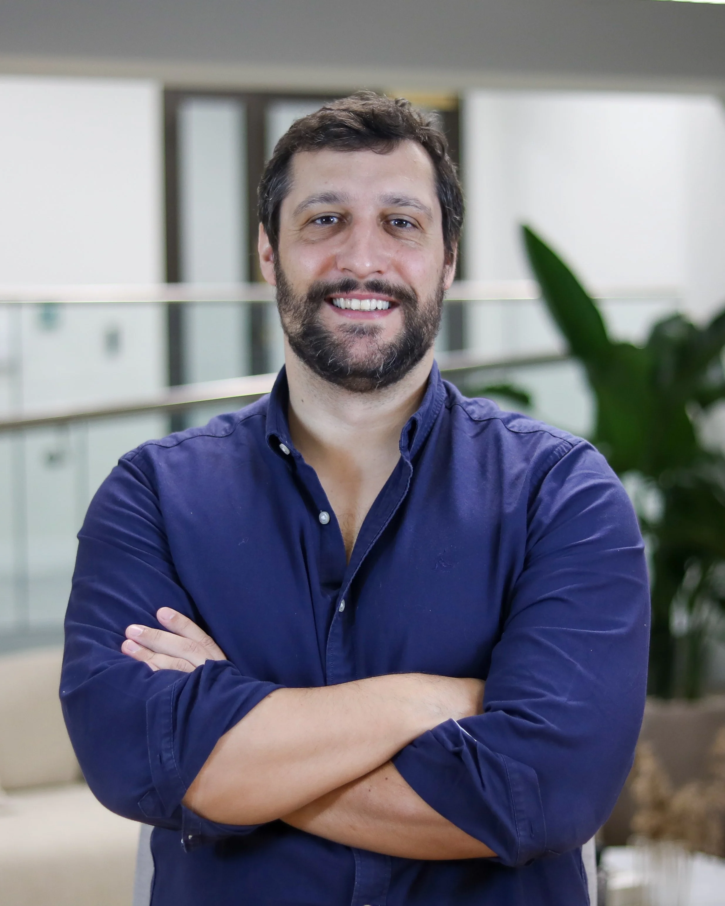 A man with brown hair and a beard, smiling with arms crossed, wearing a dark blue shirt, standing in a modern office environment.