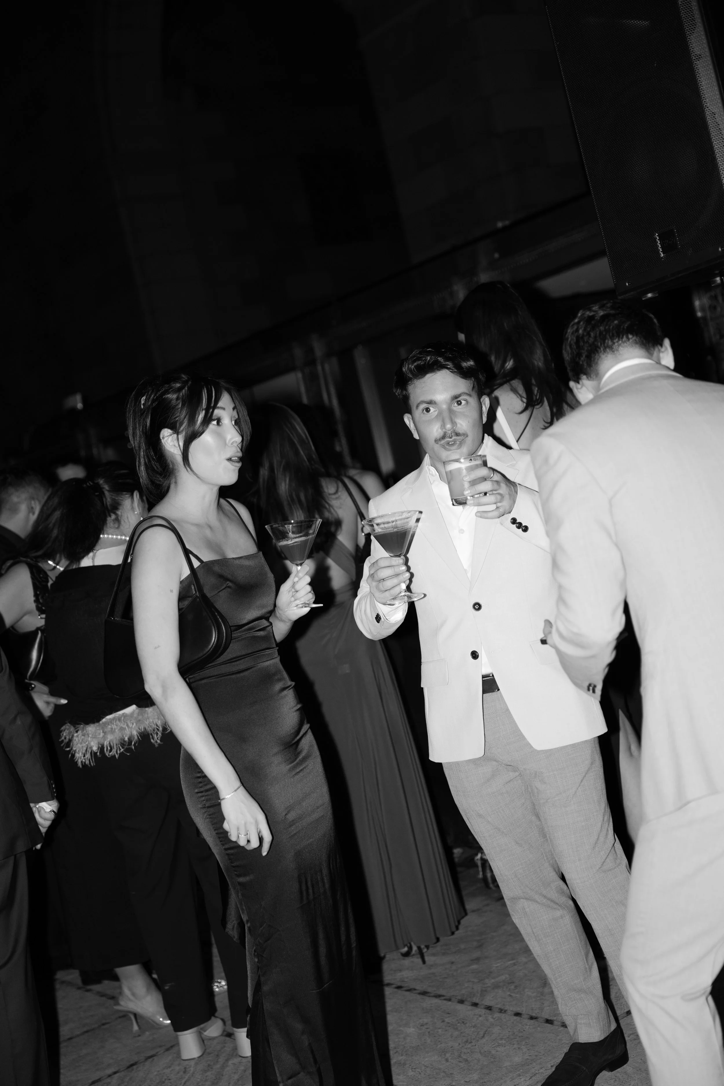 People at a social event holding drinks, engaging in conversations, dressed in formal attire, with a woman in a black dress and a purse, and men in suits, in an indoor setting.