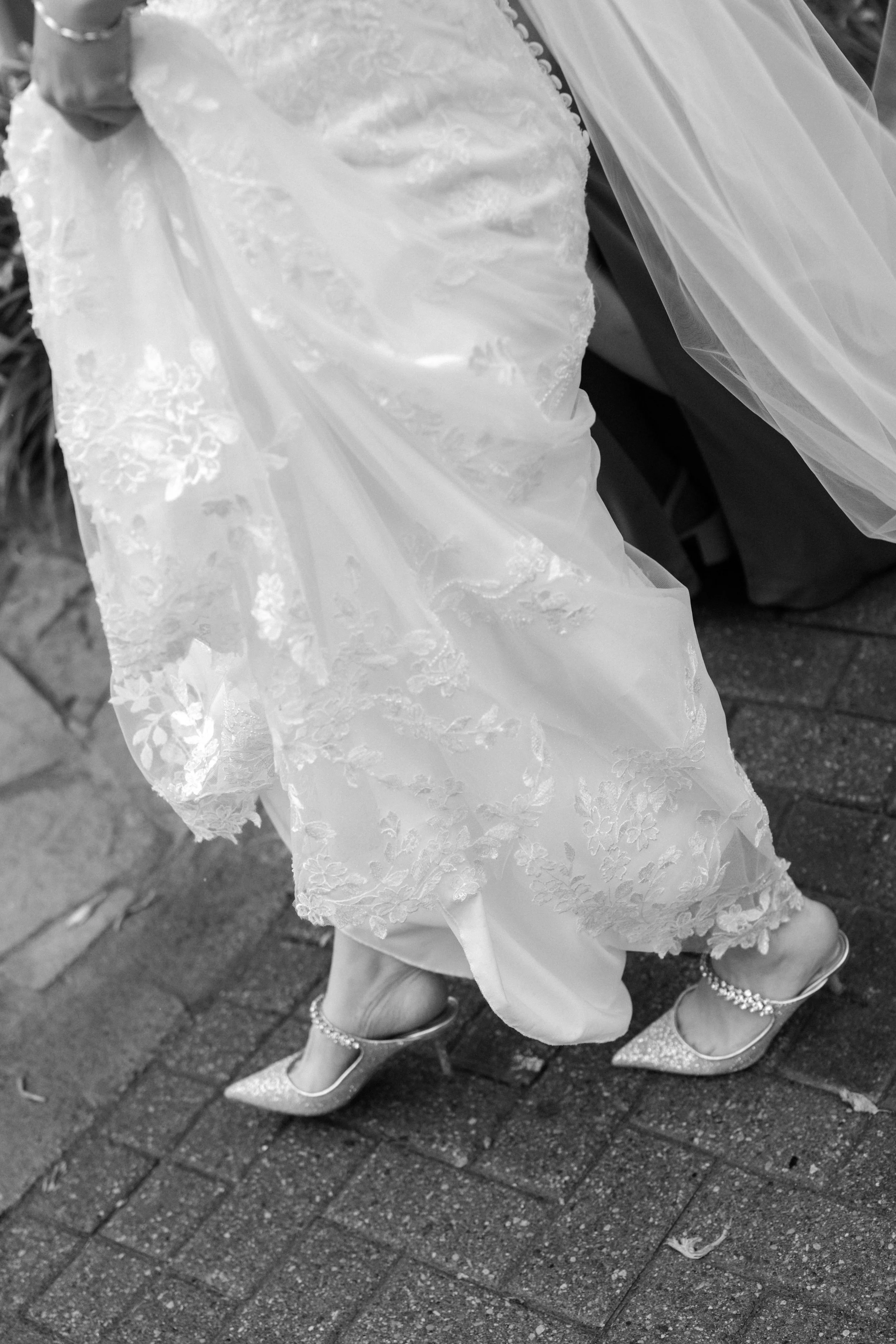 Close-up of a bride lifting her wedding dress, showing her high-heeled shoes with ankle straps and decorative embellishments, on a brick sidewalk.