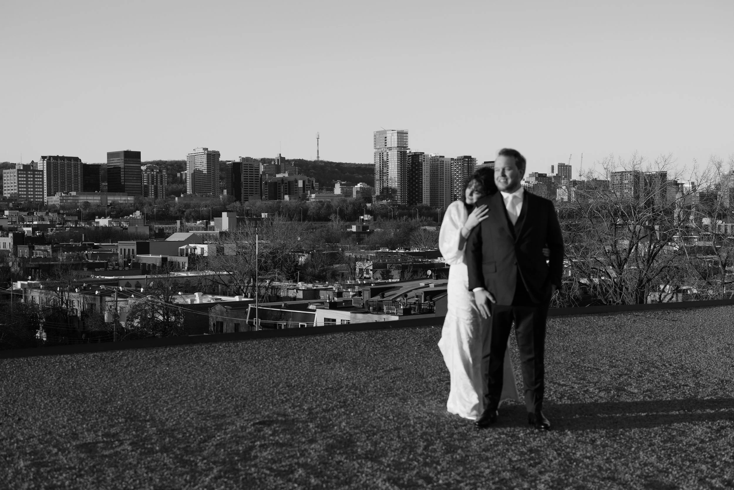 A black and white photo of a couple in wedding attire hugging on a rooftop with a city skyline in the background.