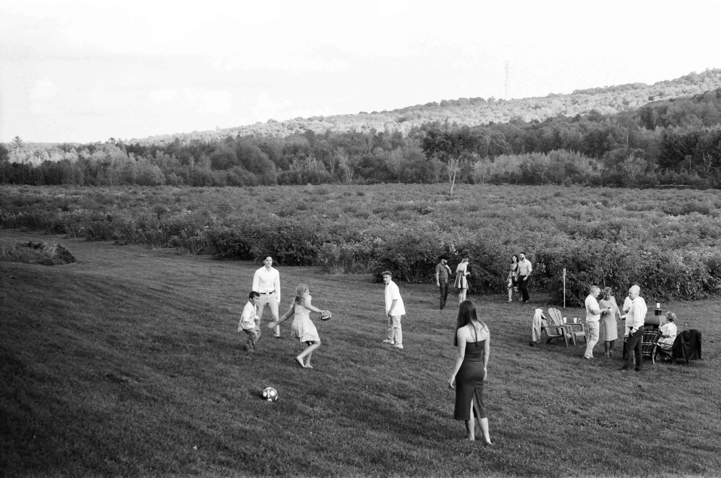 People gathered outdoors in a grassy area with a view of a forested hillside in the background. Some individuals are playing with a soccer ball, while others are standing and conversing near tables and chairs.