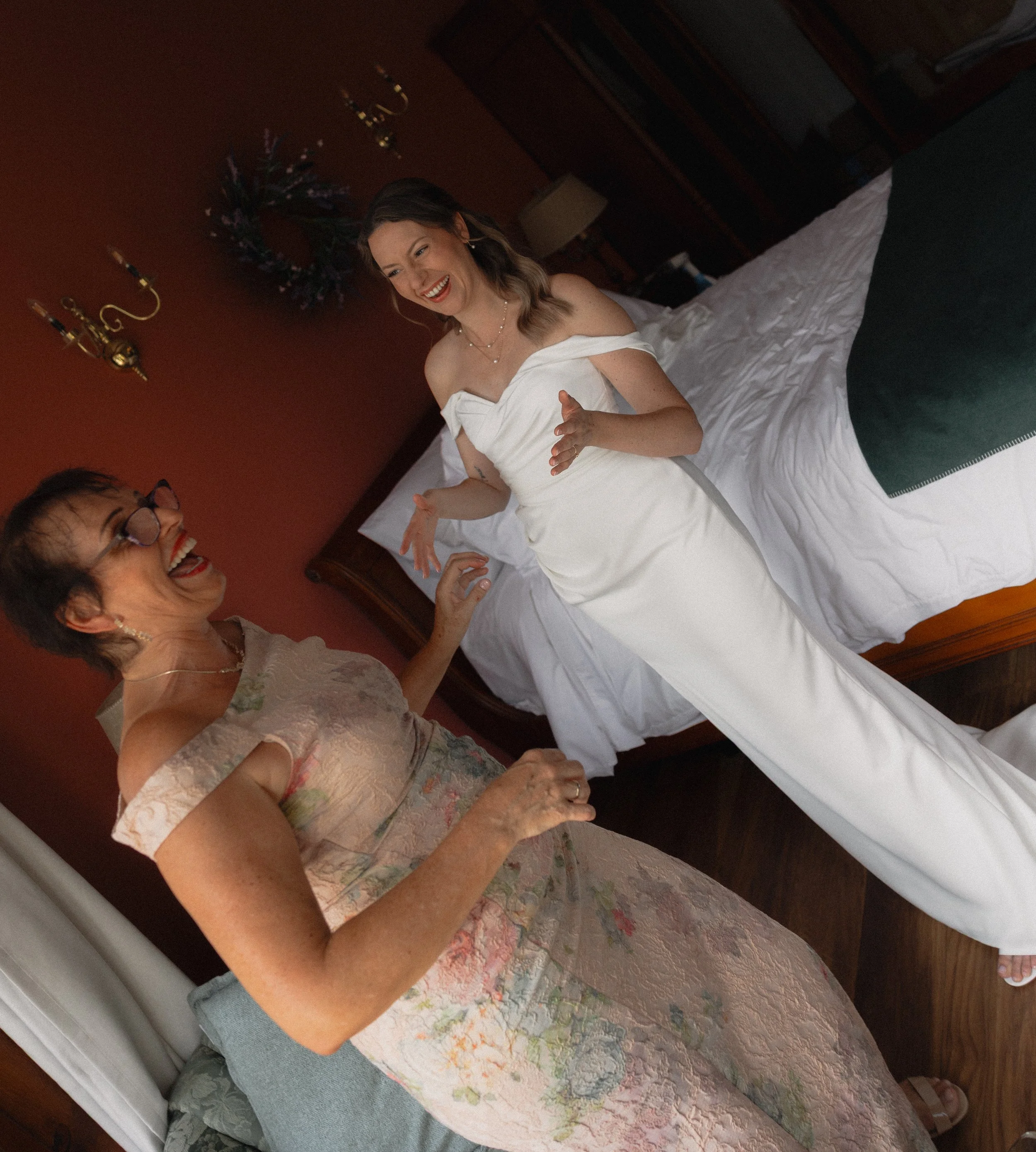 A woman in a white dress standing on a bed, smiling and laughter with an older woman in a floral dress, in a bedroom with warm lighting and dark wood furniture.