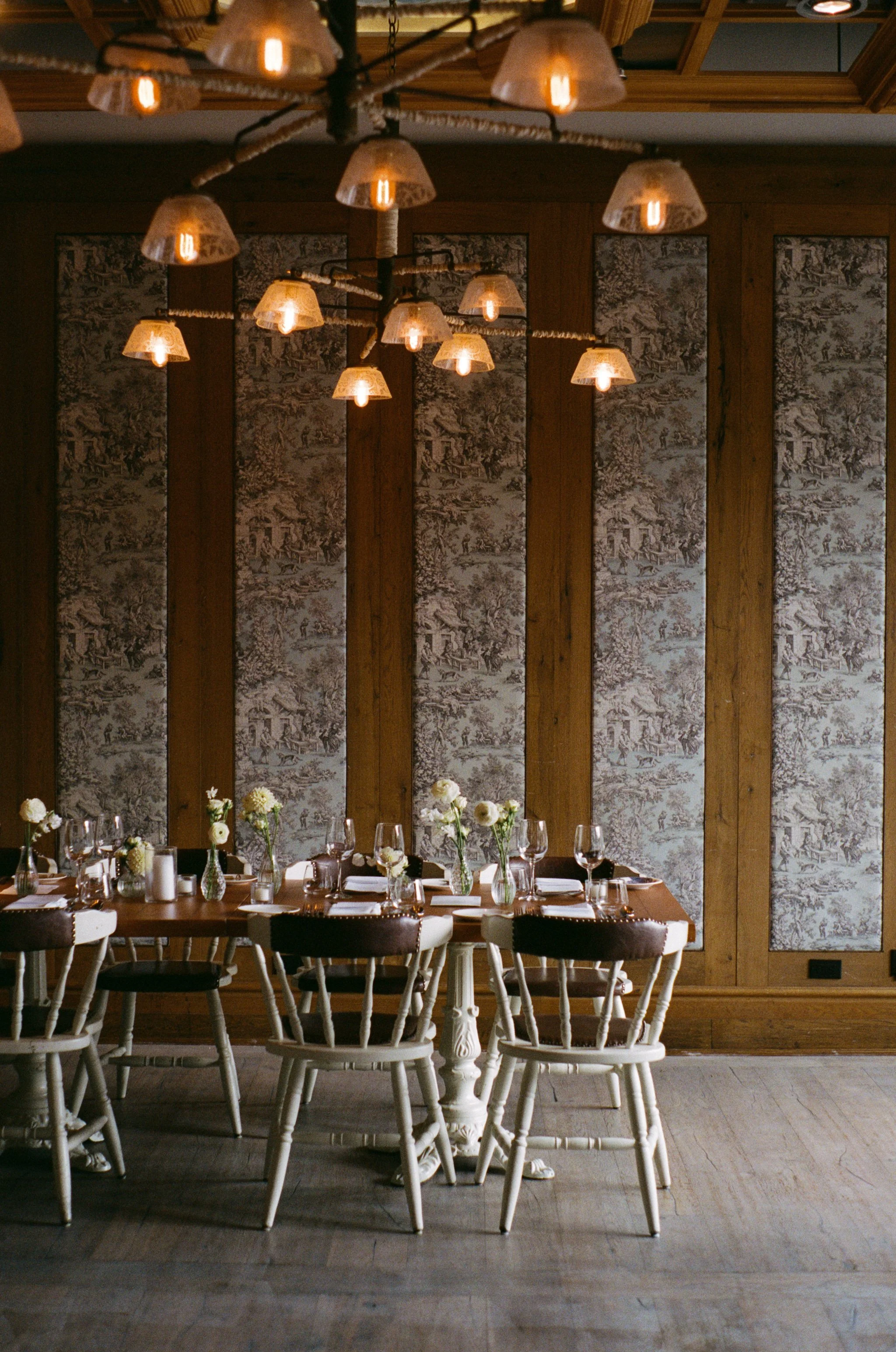 A dining room with a wooden table set with white flowers in vases, wine glasses, and tableware. The room features wood-paneled walls with intricate vintage wallpaper and a multi-bulb chandelier with decorative covers hanging from the ceiling.