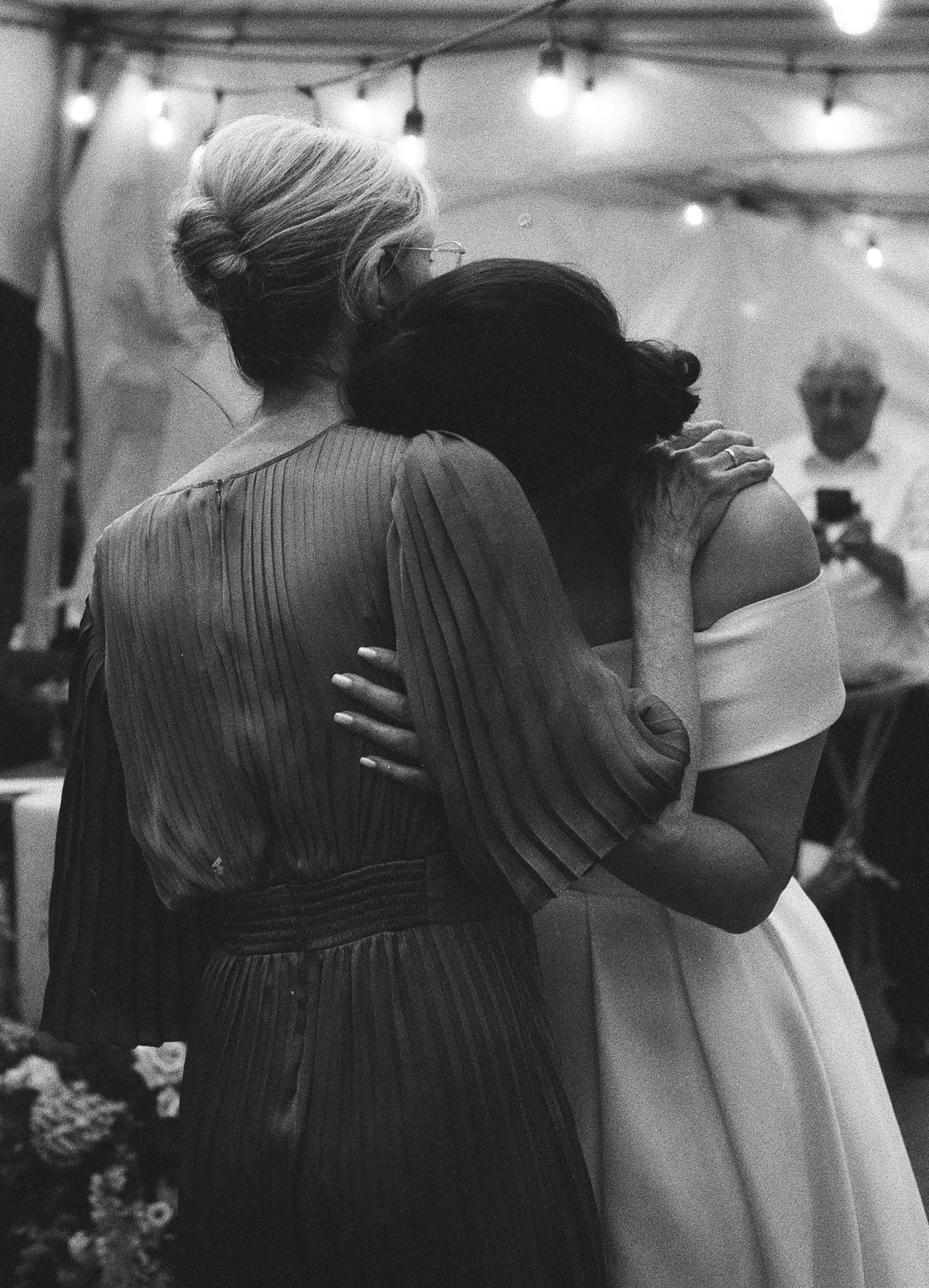 Two women embrace each other in a heartfelt hug, one in a dark dress and the other in a white off-the-shoulder dress, inside a setting decorated with string lights, with a man in the background taking a photo.