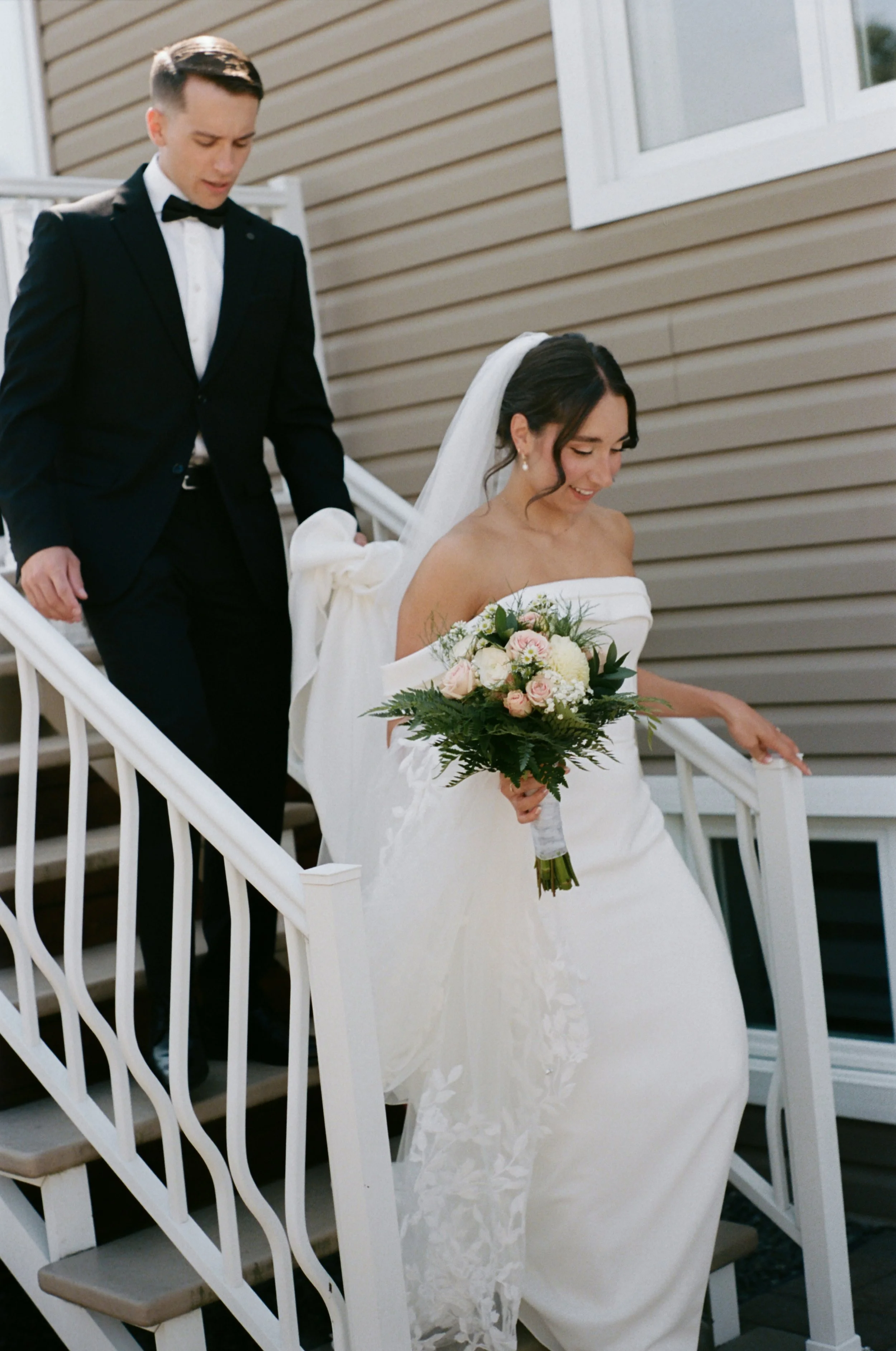 Bride in a white wedding gown with a bouquet ascending a staircase, groom in a black tuxedo.