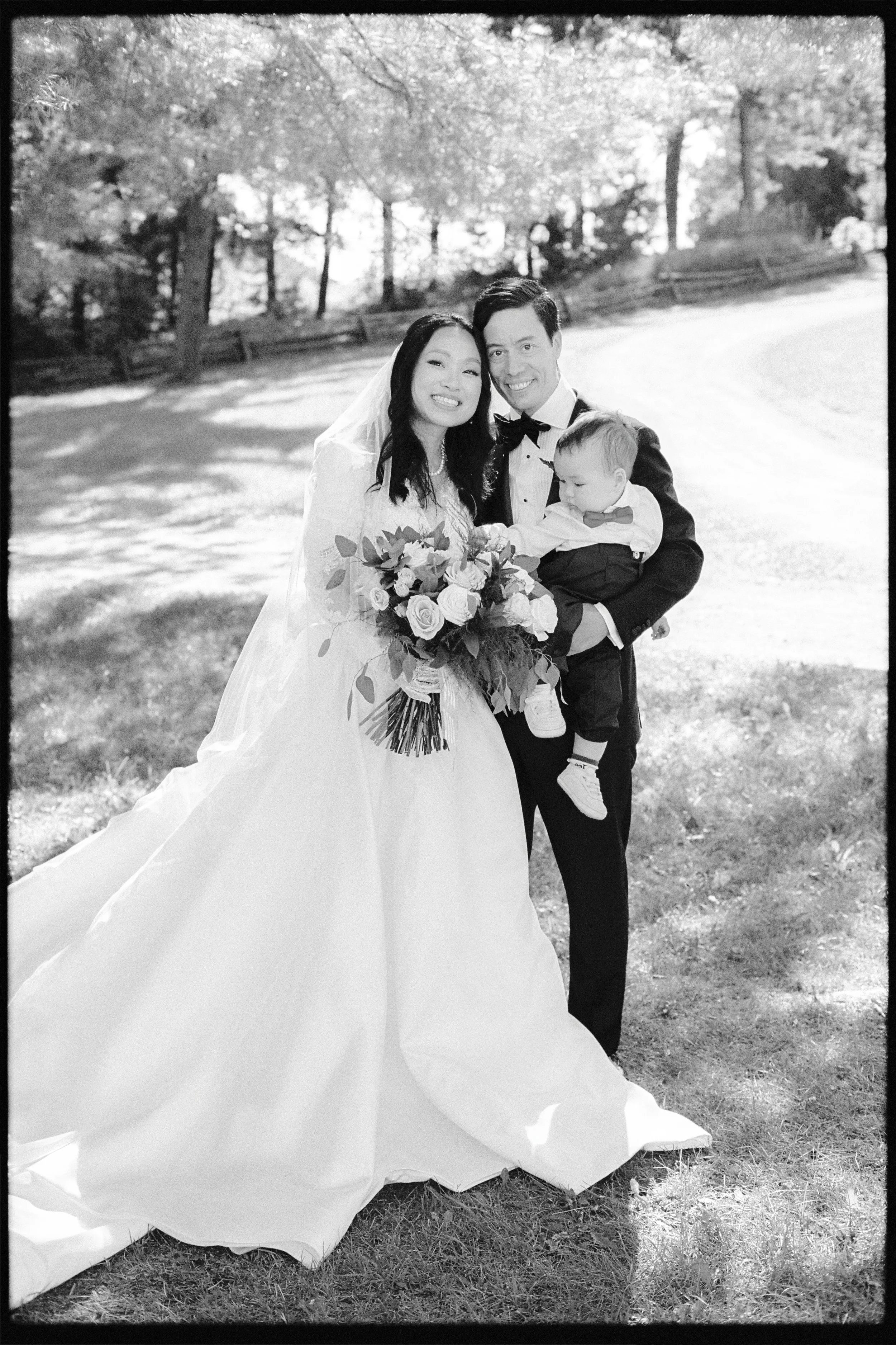 A wedding photograph in black and white showing a bride and groom smiling and holding a child outdoors in a park with trees in the background. The bride is in a wedding dress with a veil and holding a bouquet of flowers. The groom is in a tuxedo, and
