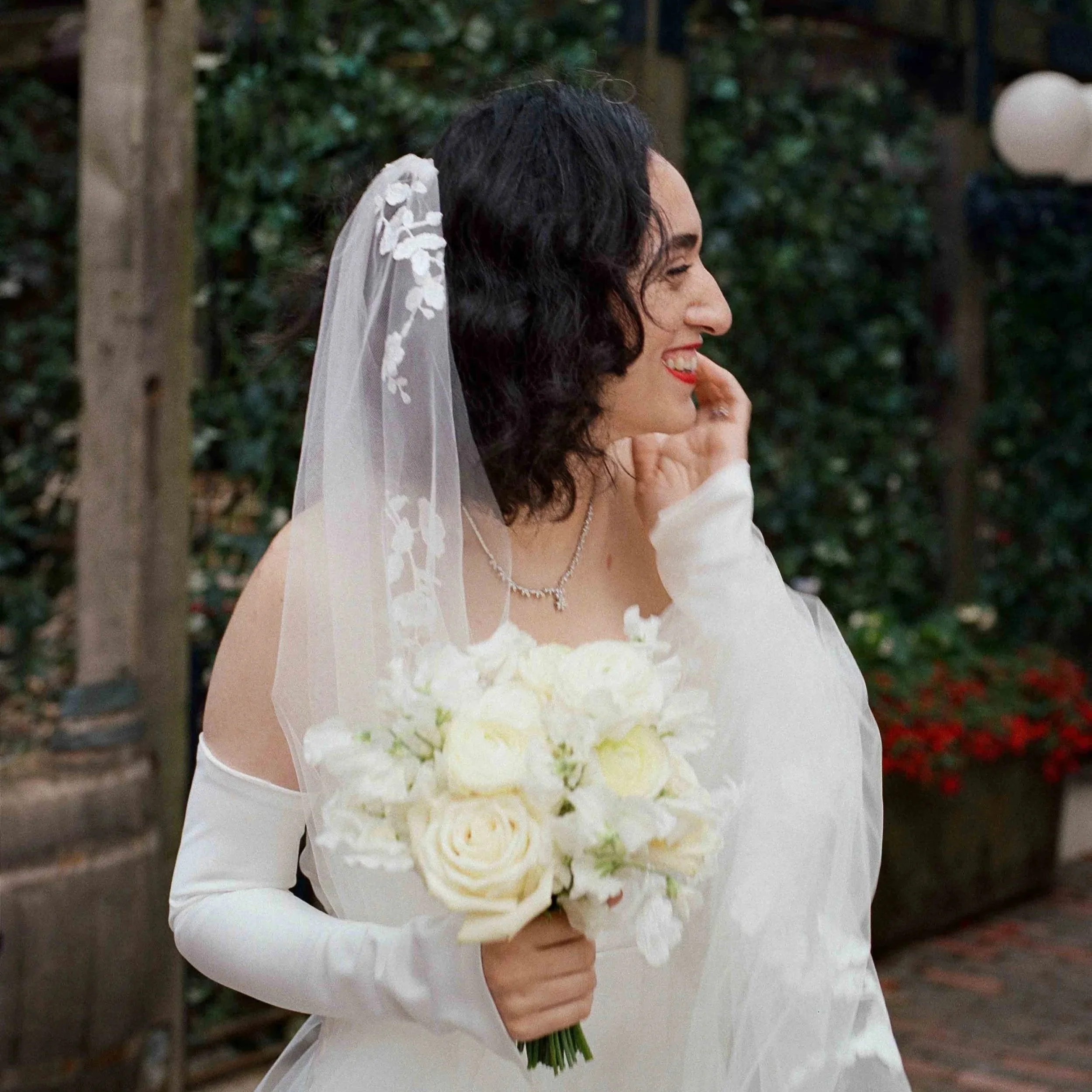 A woman in a white dress holding a bouquet of white roses, with a veil on her head, smiling and looking to her right outdoors with trees and wall as background.