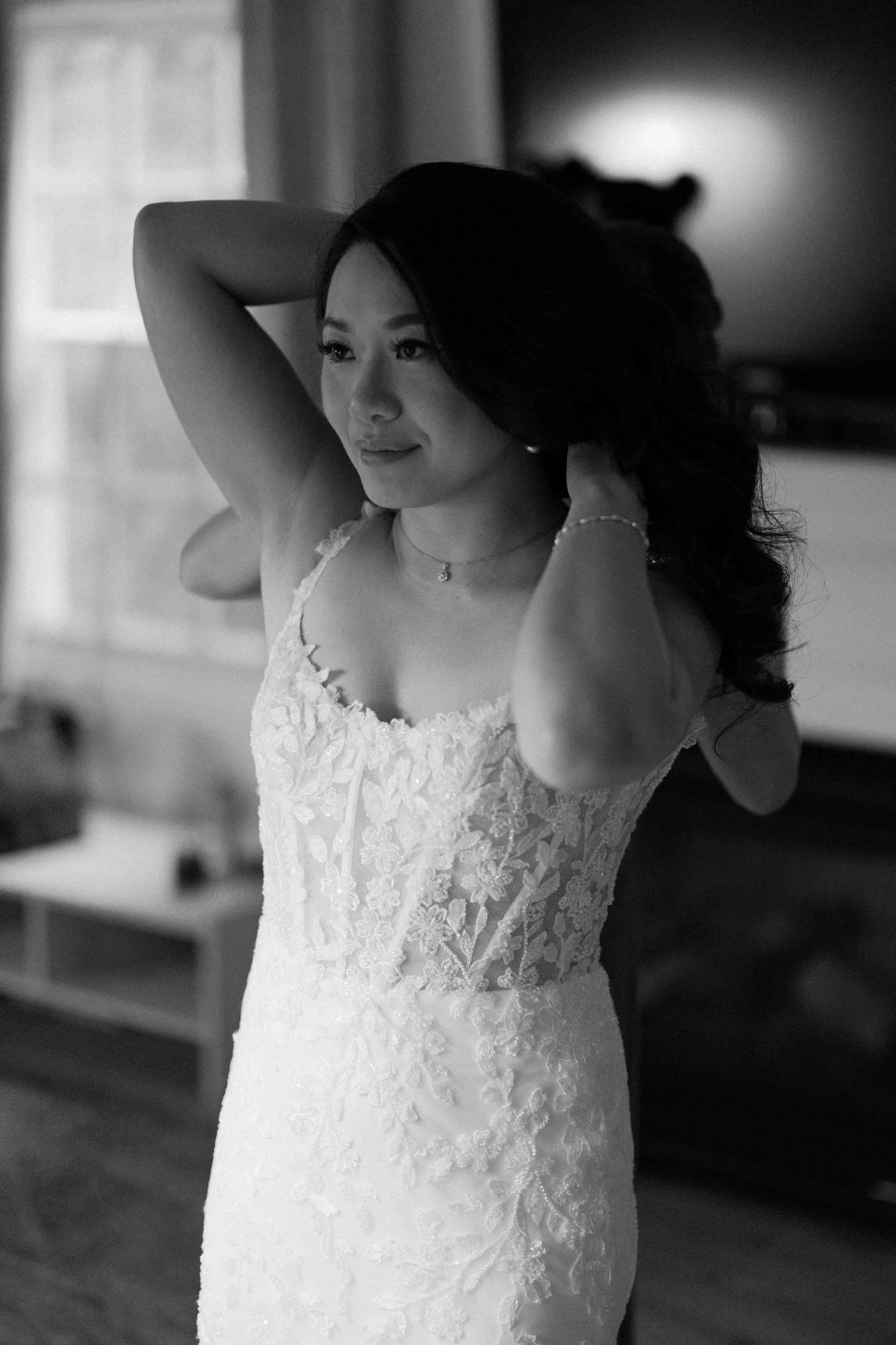 A woman in a white lace wedding dress, adjusting her hair, standing indoors in front of a window with curtains.