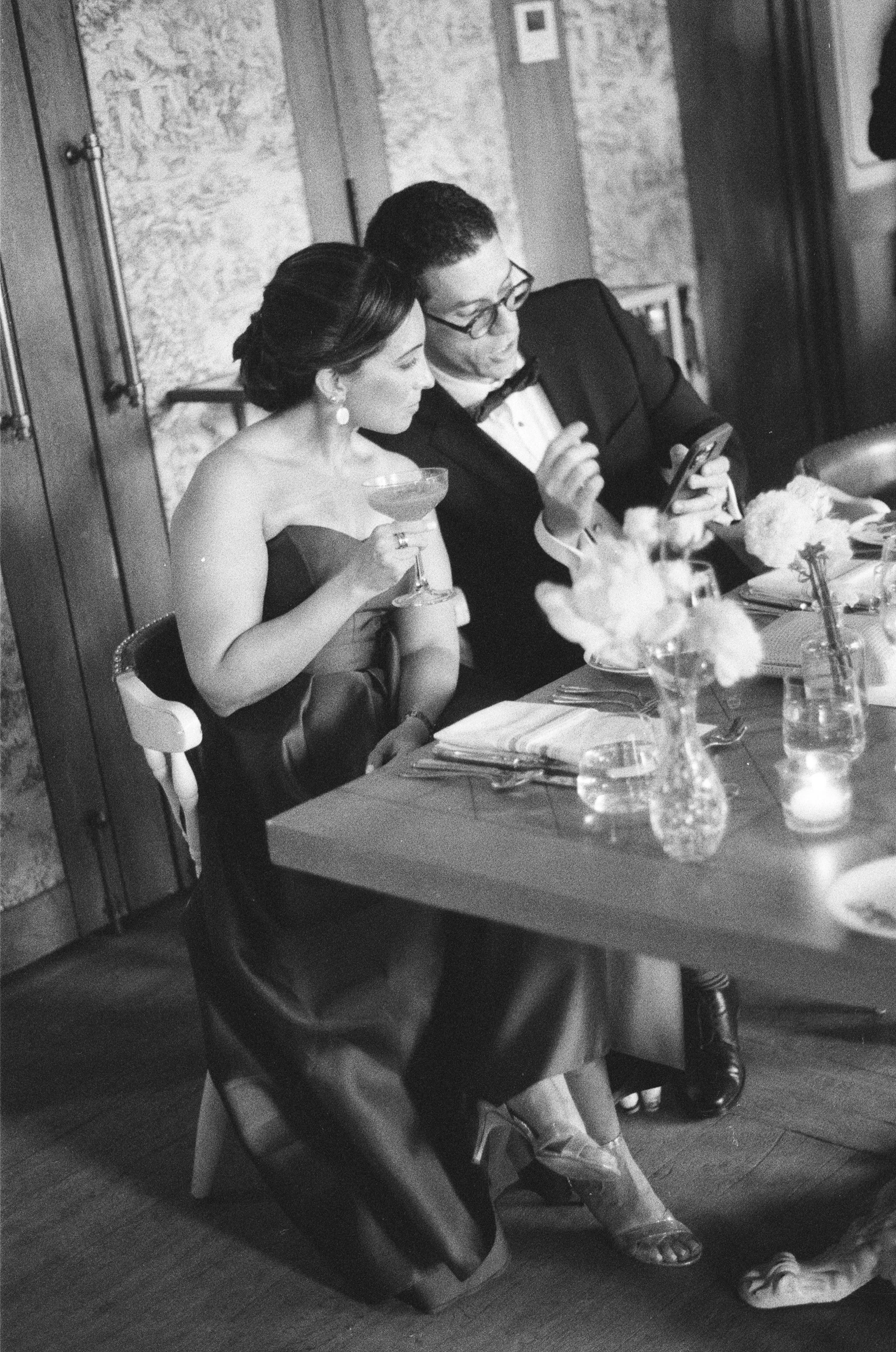 A couple in formal attire at a dinner table, the woman is holding a cocktail glass, and the man is looking at his phone.