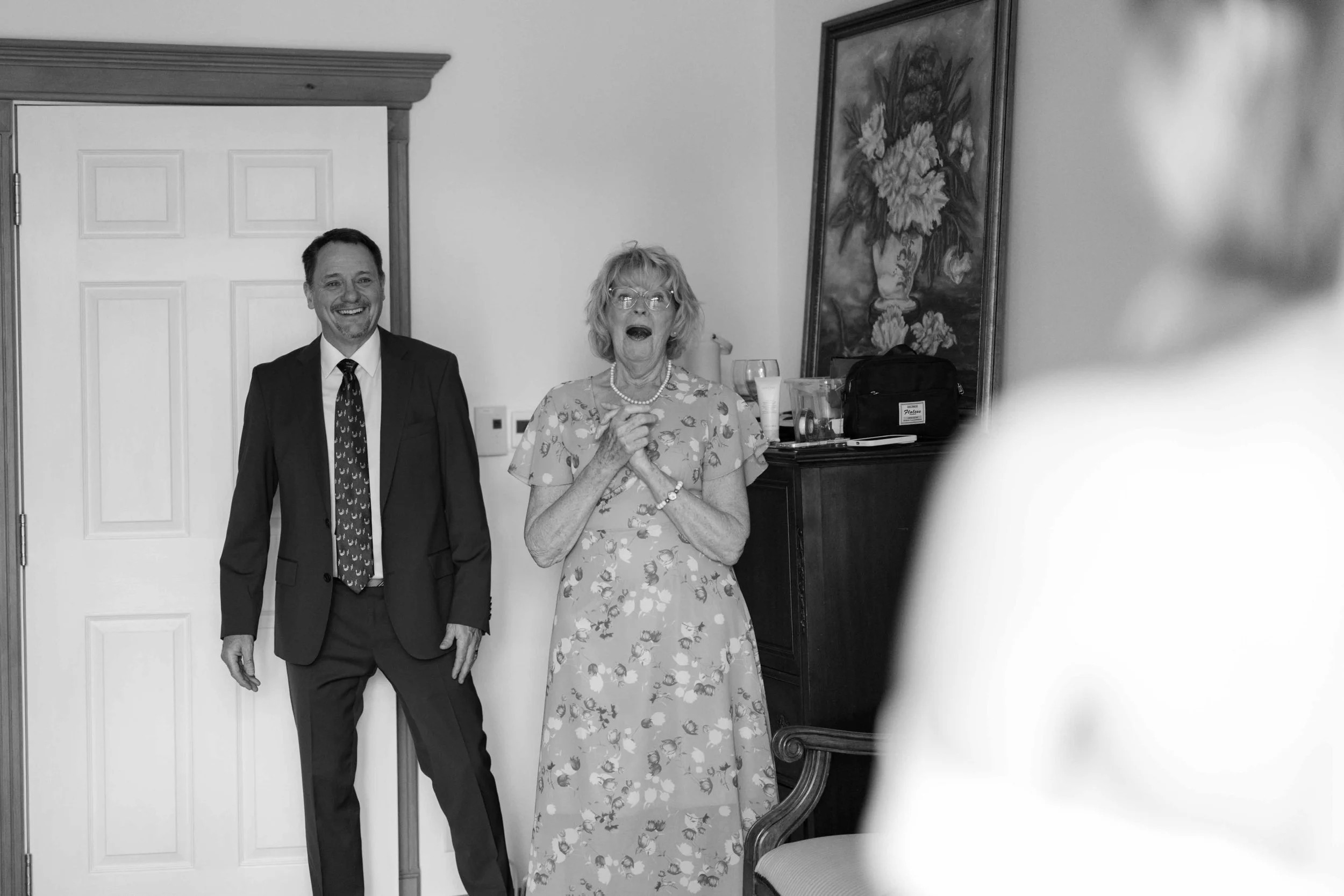 A black and white photo showing a man in a suit and a woman in a floral dress standing in a room with a door and a painting of flowers on the wall.