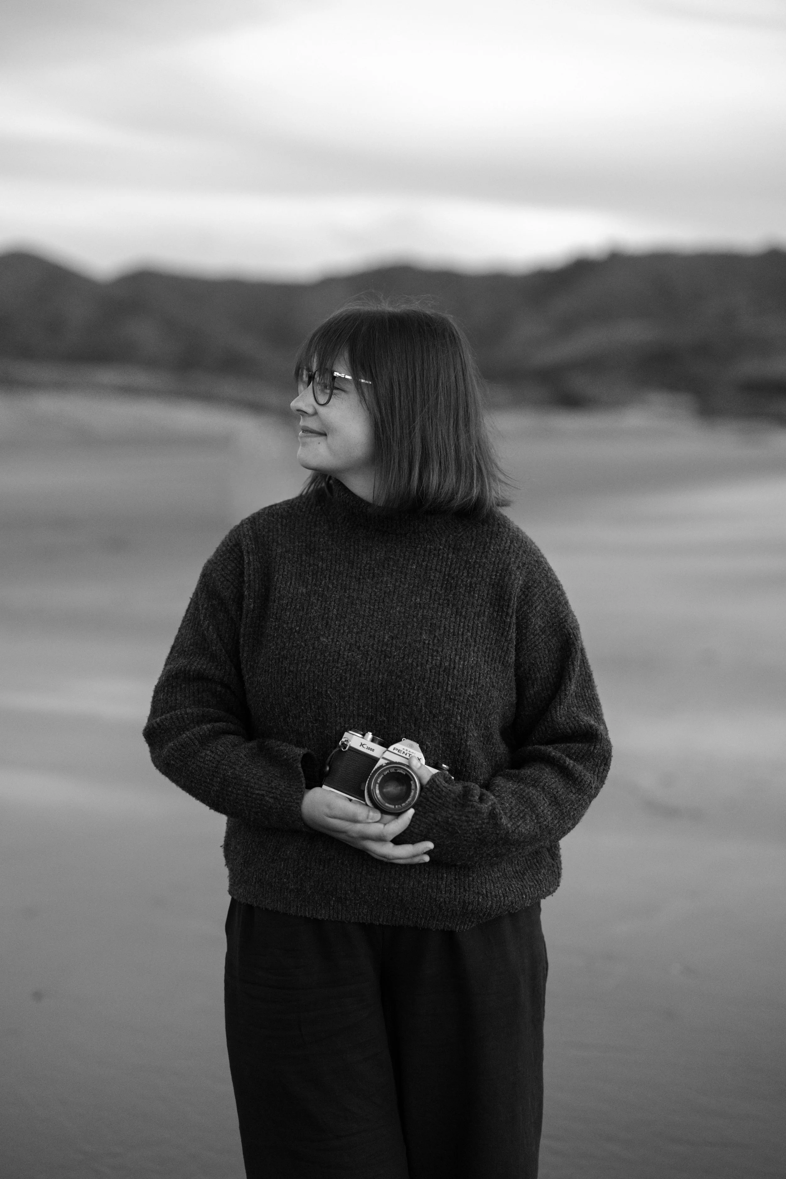 A woman with glasses holding a camera stands outdoors on a beach, with hills and cloudy sky in the background.