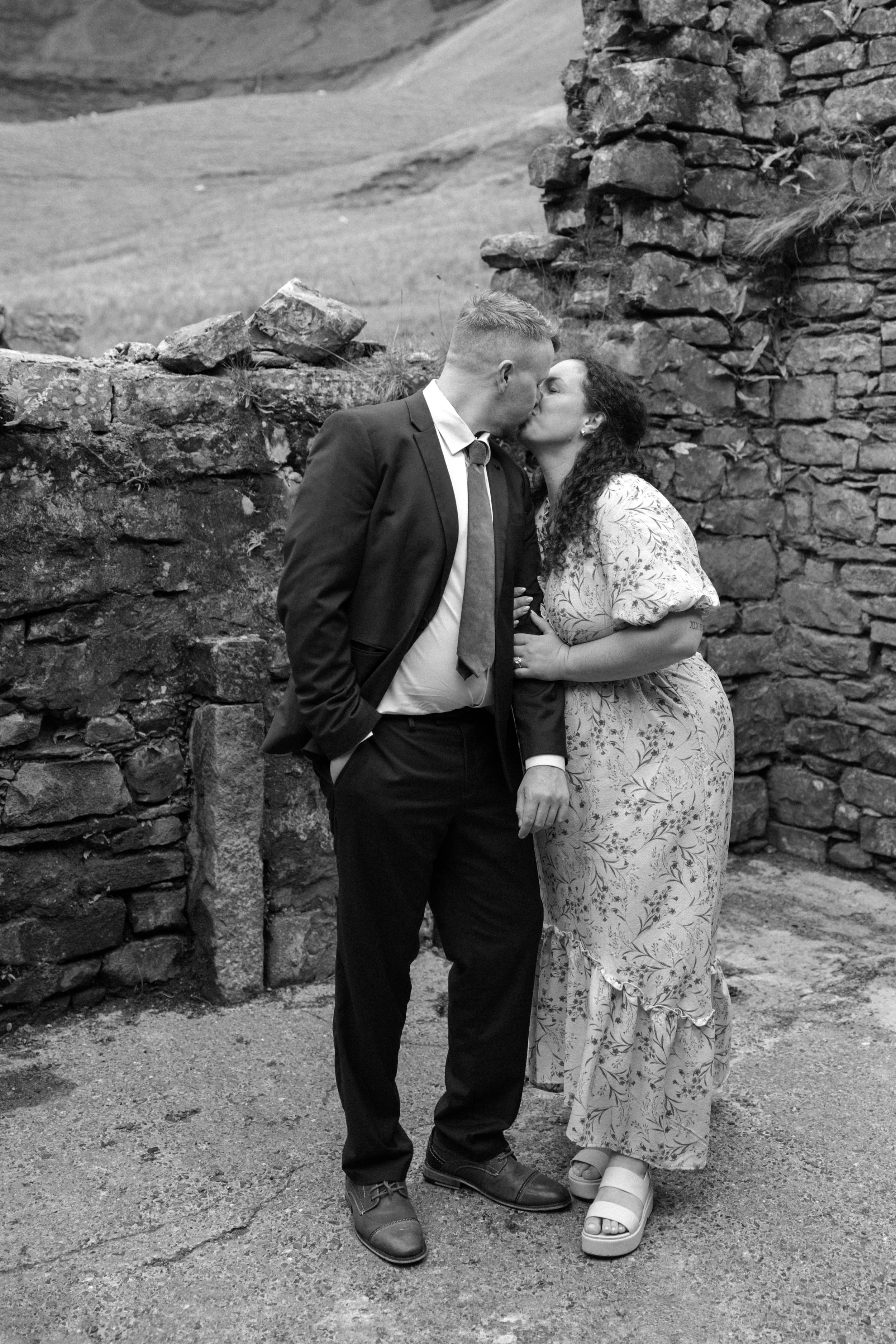 A couple in formal attire sharing a kiss outdoors, standing near a stone wall.