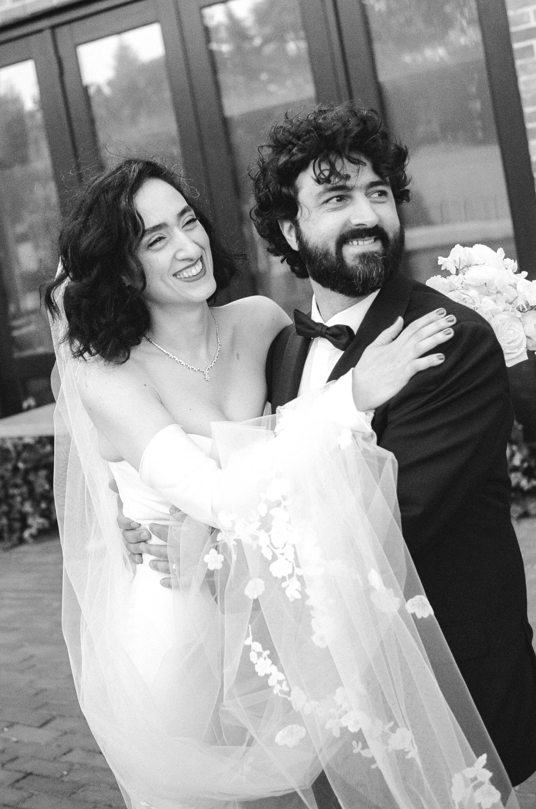 Black and white photo of a bride and groom smiling, with the bride wearing a strapless wedding gown and veil, and the groom in a tuxedo with a bow tie, outside near a glass building.