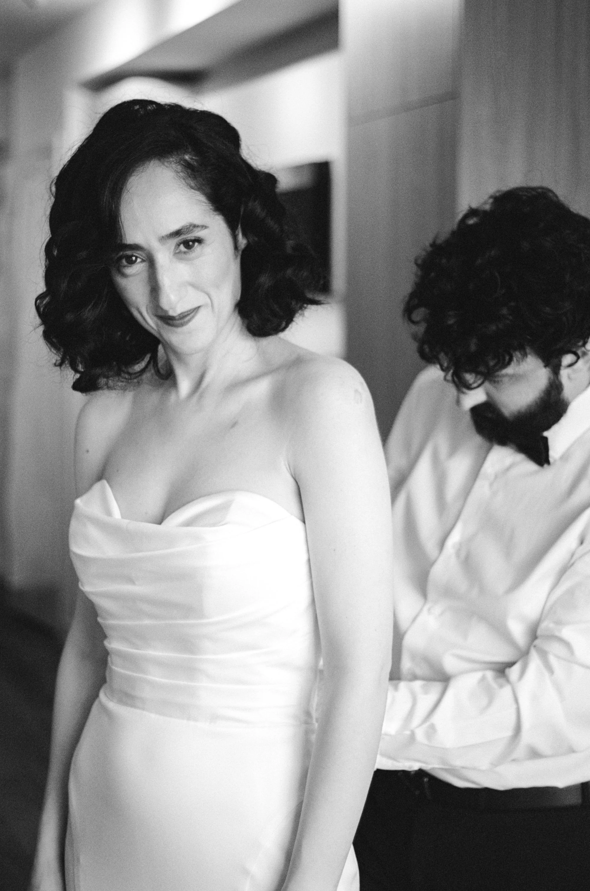 A woman in a white strapless dress with dark, wavy hair smiling at the camera, while a man with curly hair and glasses adjusts her dress in a black and white photo.