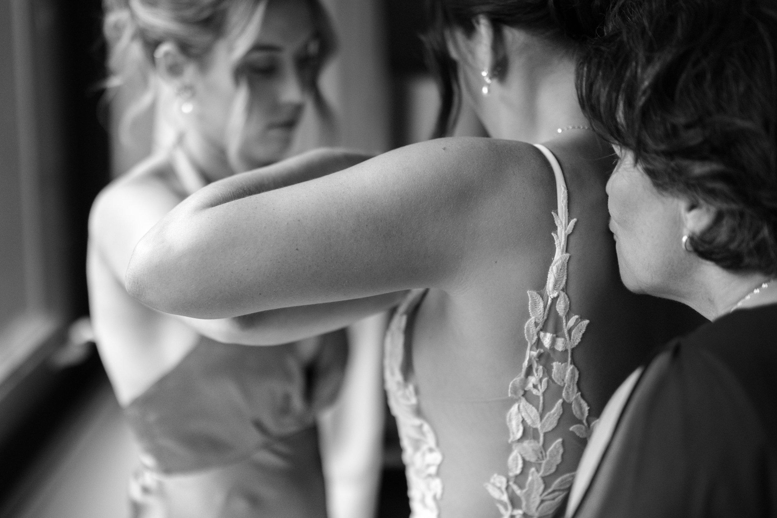 Black and white photo of a young woman getting ready, with another woman helping her with her dress, both smiling and focused on the garment.