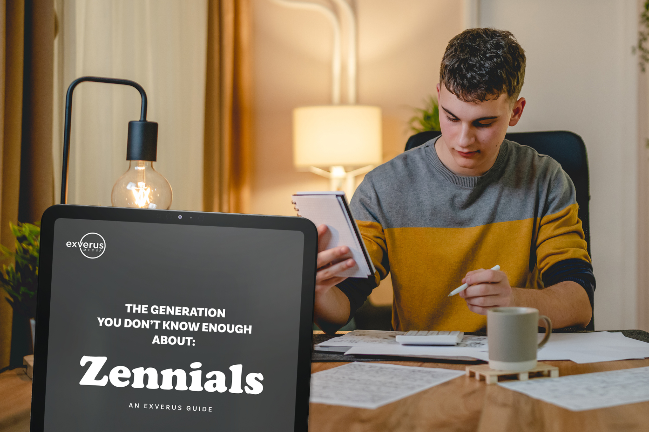 A young man working at a desk with a laptop, notepad, and documents, in a warmly lit room with a table lamp and curtains.