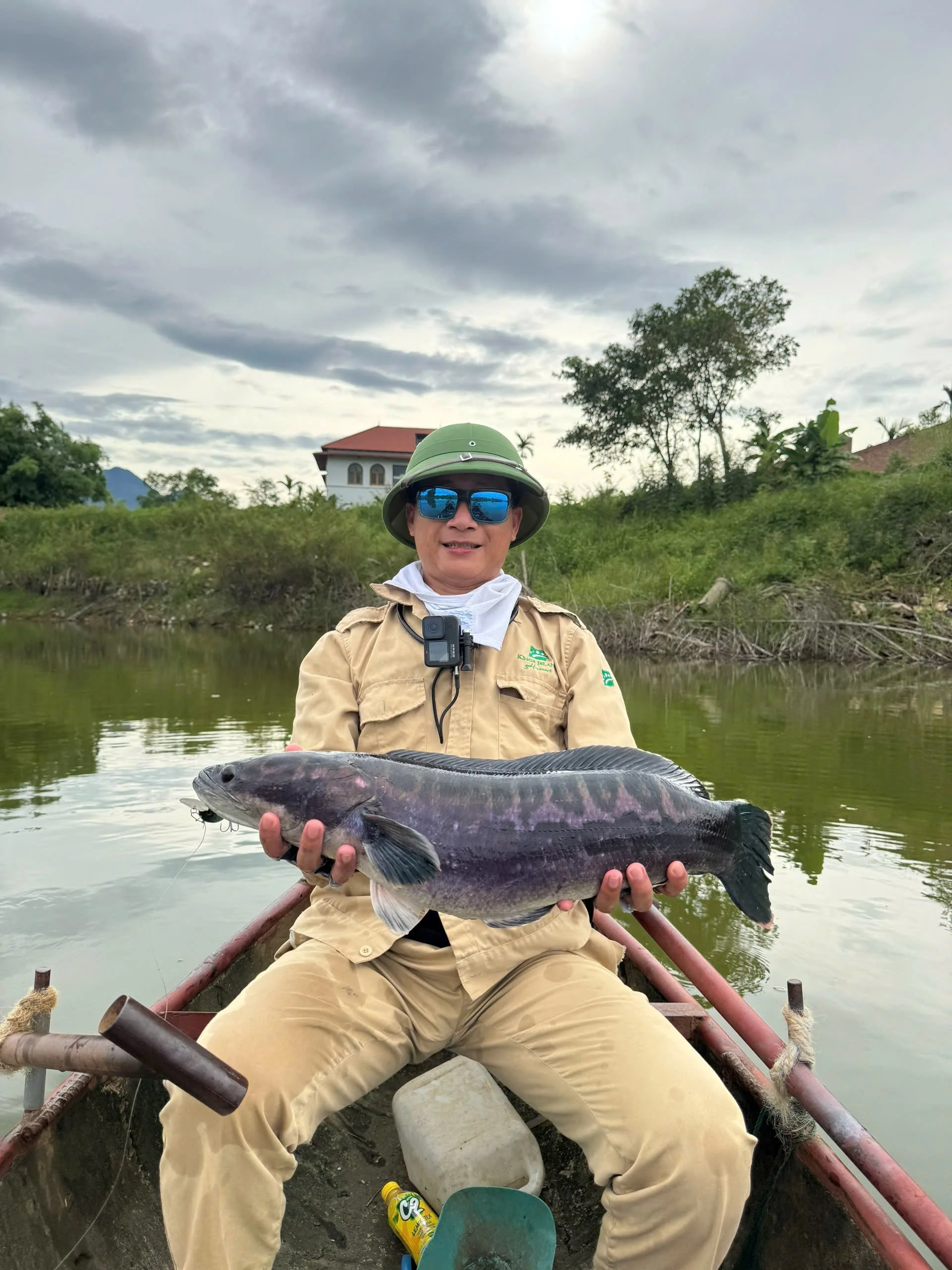 A person sitting in a small boat on a river, holding a large fish, wearing sunglasses, a green hat, and beige outdoor gear, with a house and trees in the background.