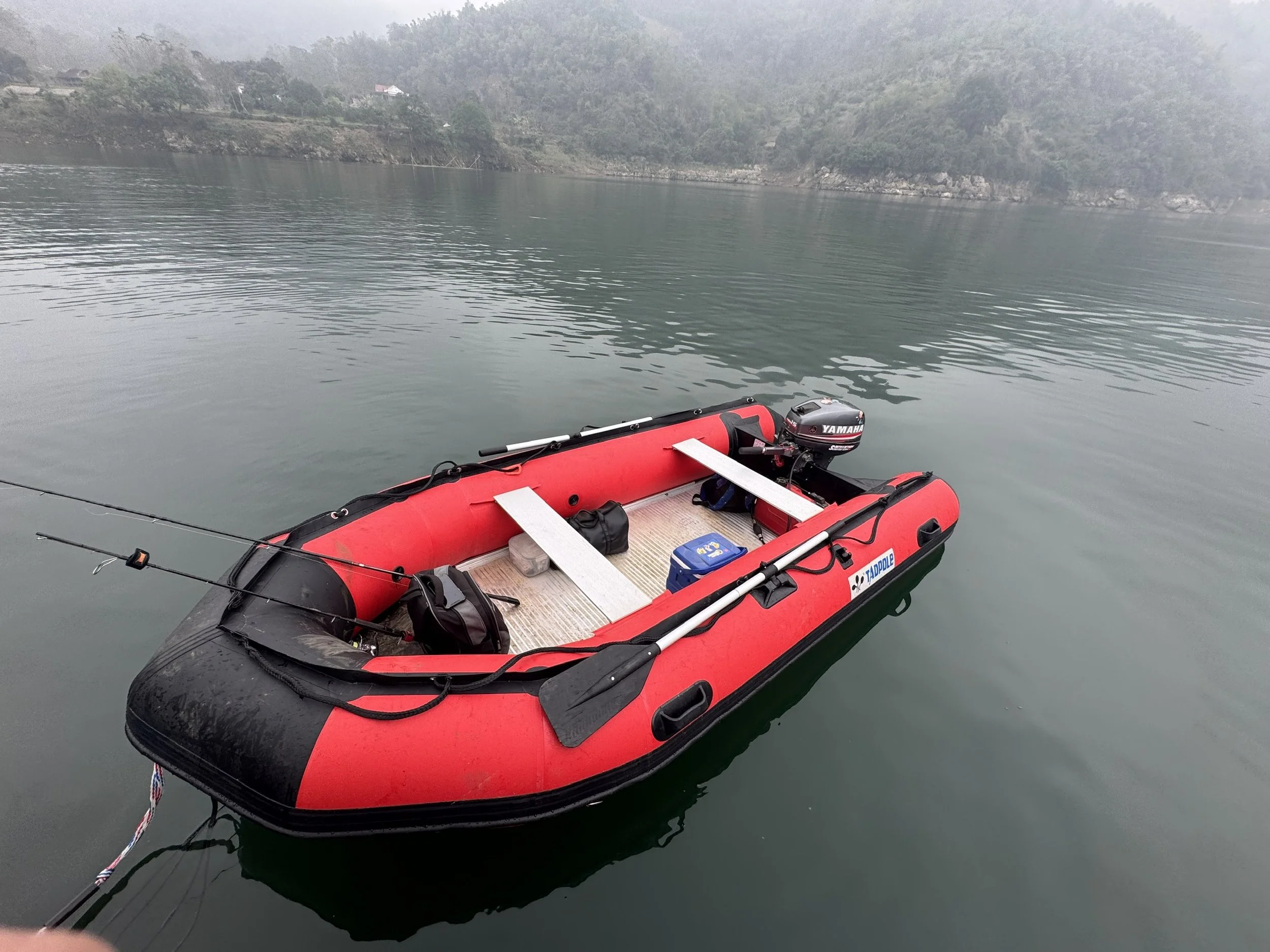 A red inflatable boat with black accents floating on calm water, equipped with a Yamaha outboard motor and fishing rods, with distant hills and a foggy sky in the background.