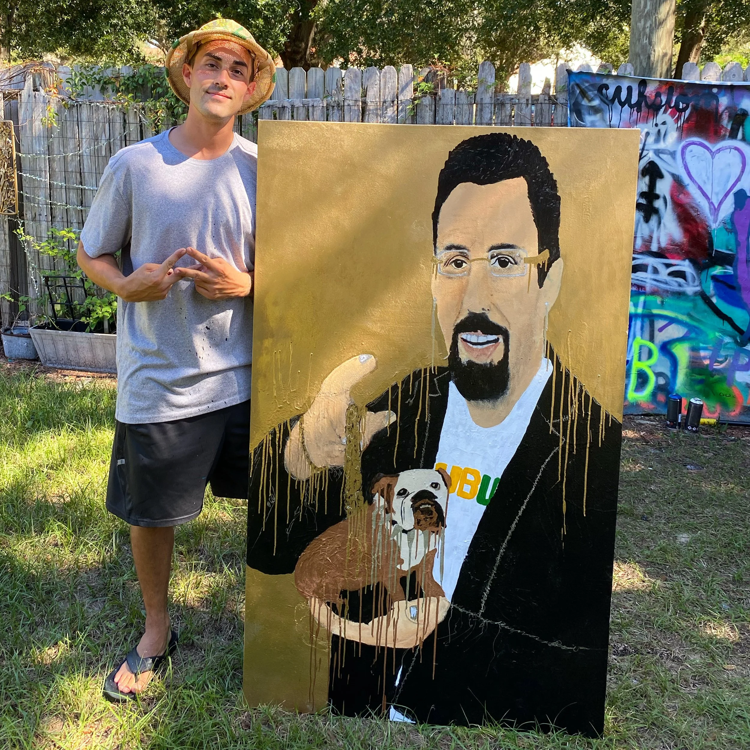 A man stands outdoors next to a large painting of a man with glasses and a goatee, holding a small dog with a similar face. The painter is wearing a straw hat, a light gray t-shirt, black shorts, and flip-flops. The background shows a wooden fence, t