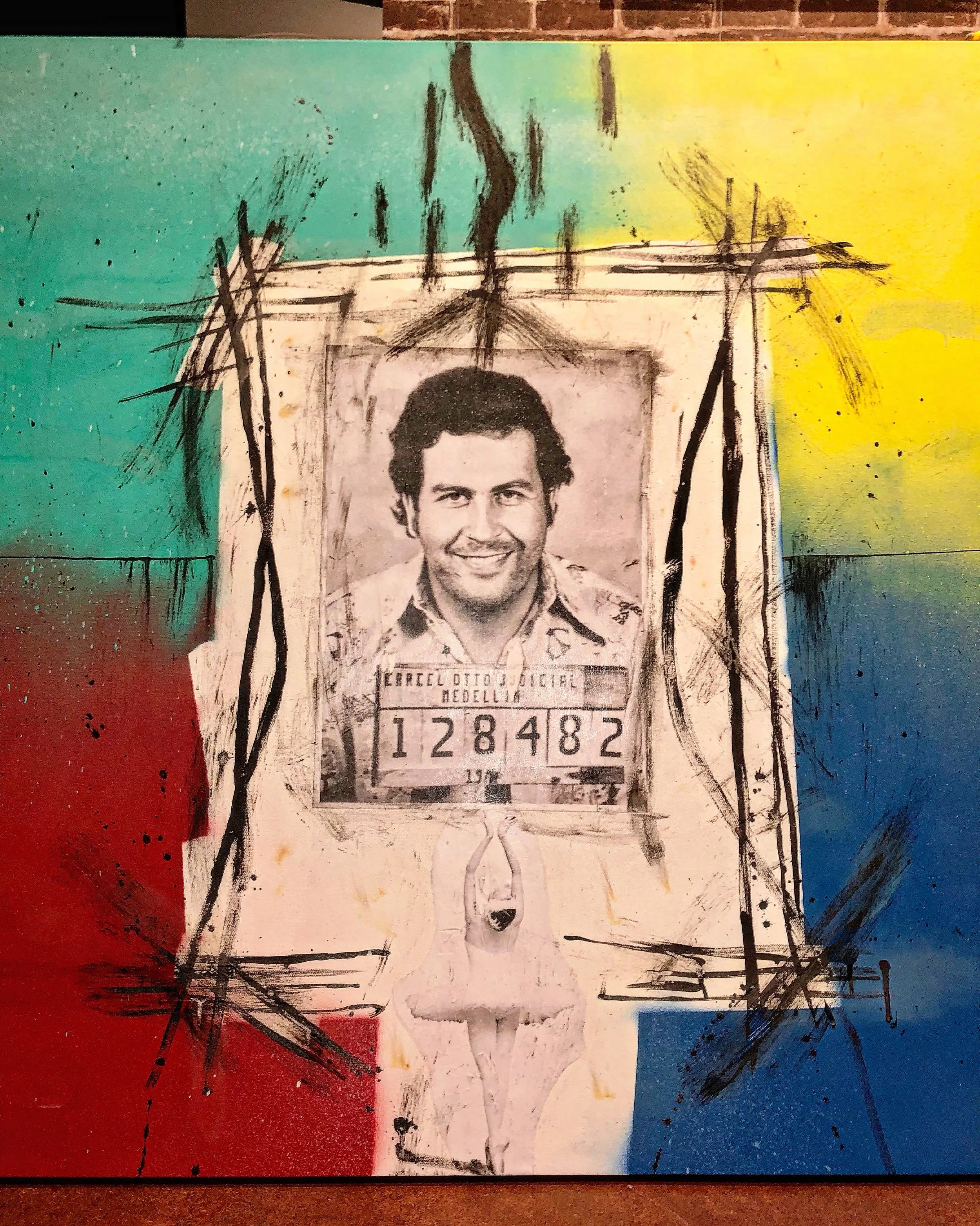 Mixed media art piece featuring a black and white photograph of a man with a smiling face, arched eyebrows, and curly hair, holding a sign with the number '128482'. The background is painted in vibrant red, blue, green, yellow, and black with abstrac
