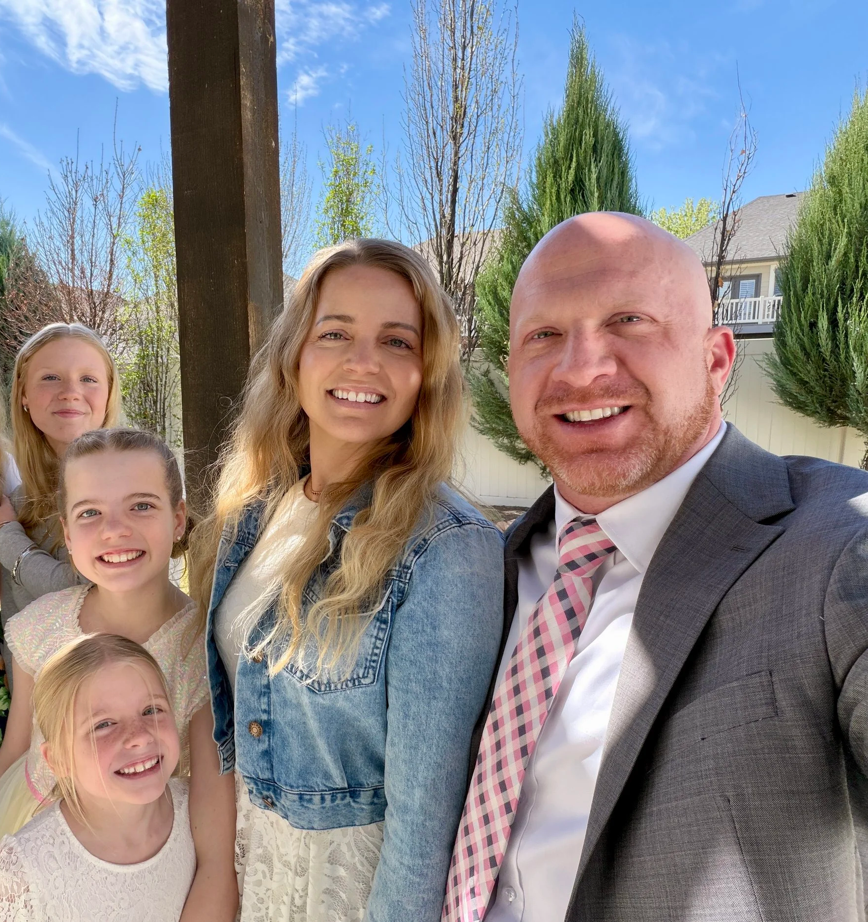 A group of smiling children and adults outdoors on a sunny day, with trees and a house in the background.
