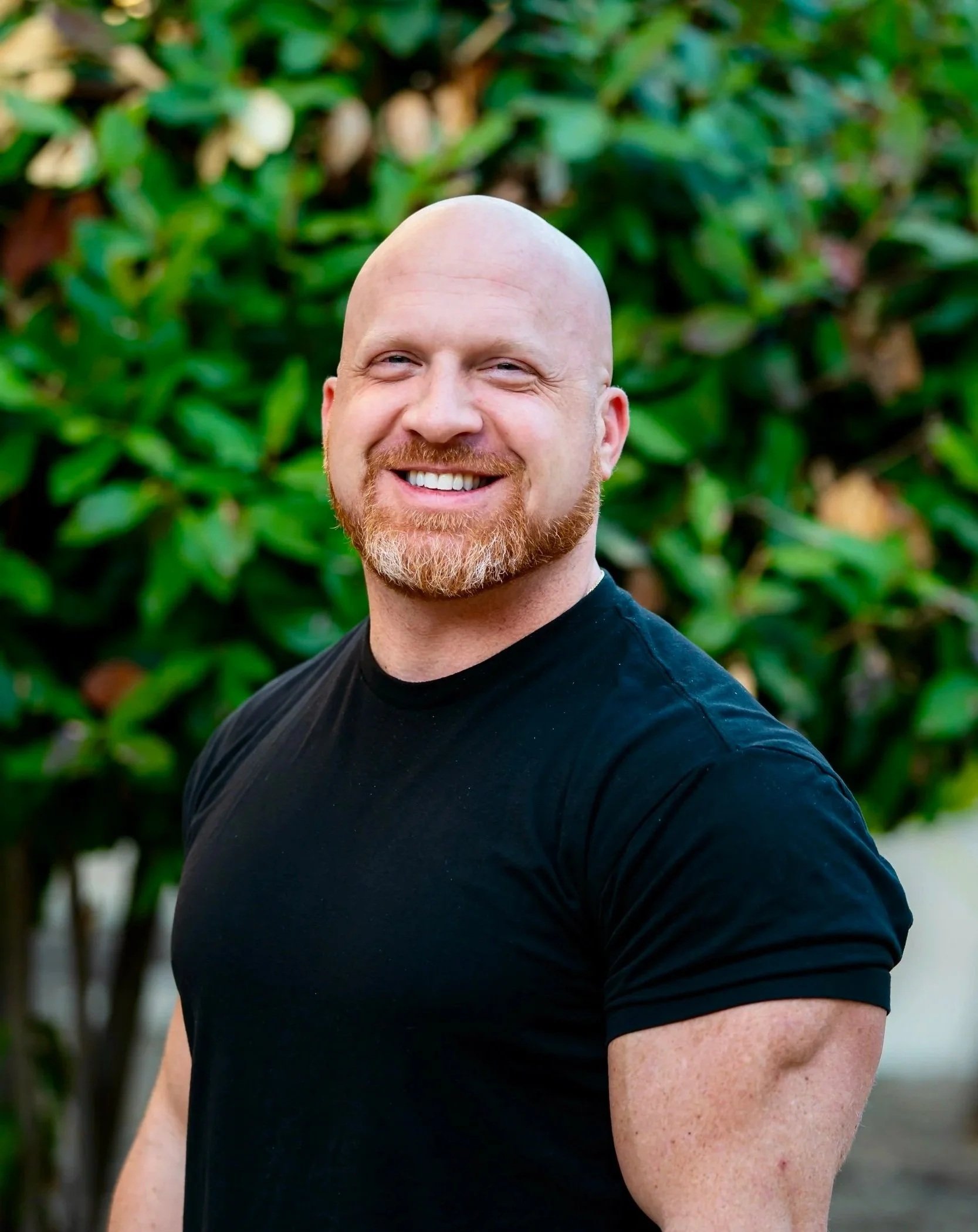 Close-up of a smiling bald man with a beard wearing a black T-shirt outdoors with green foliage in the background.