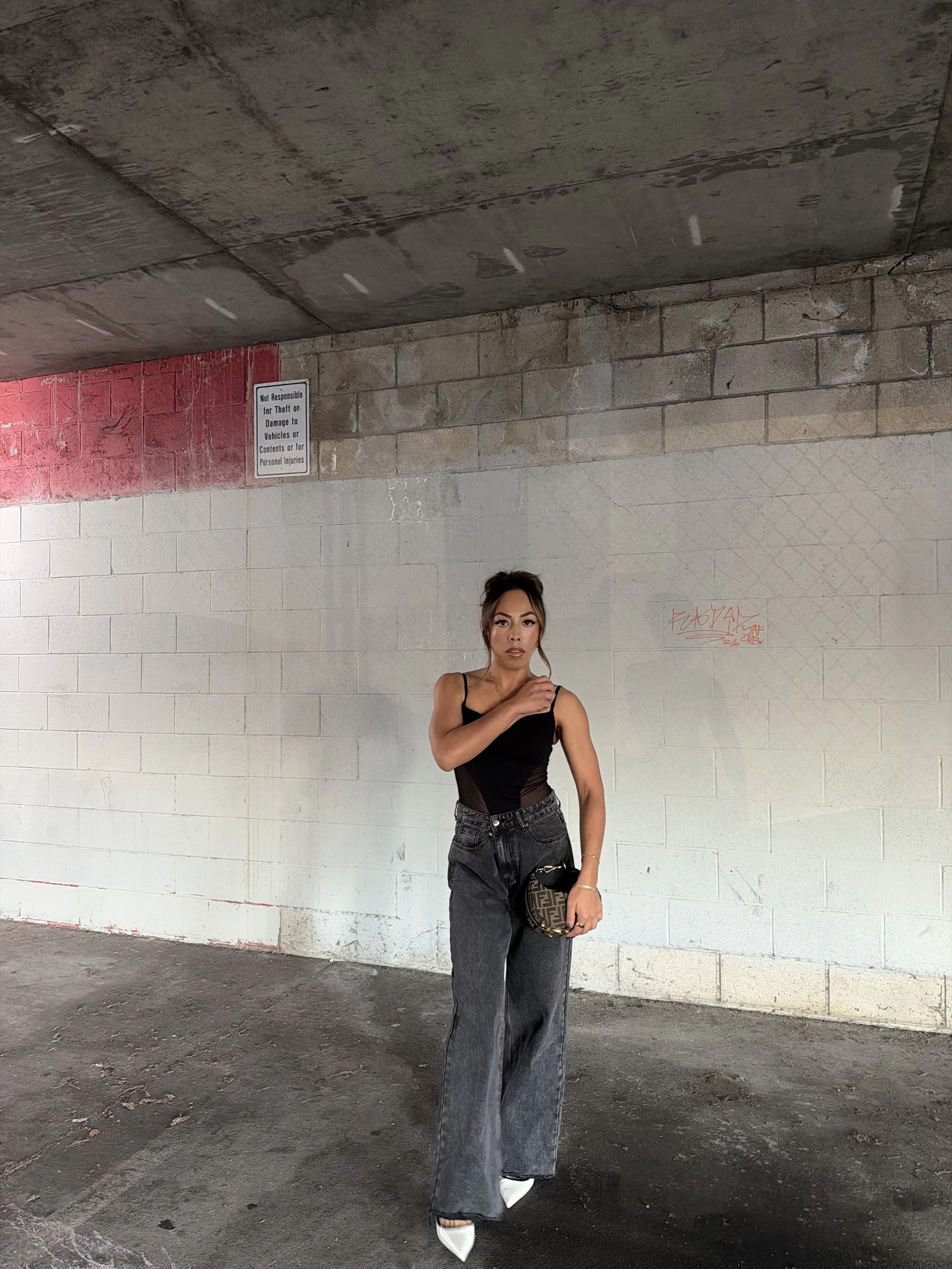 A woman with brown hair styled in an updo, wearing a black sleeveless top, high-waisted black jeans, white pointed-toe shoes, and carrying a designer handbag, standing against a concrete wall in an indoor parking area.