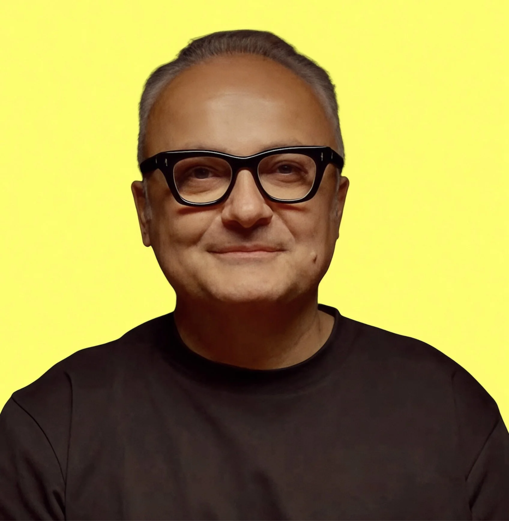 A middle-aged man with glasses, wearing a black shirt, smiling, against a bright yellow background.