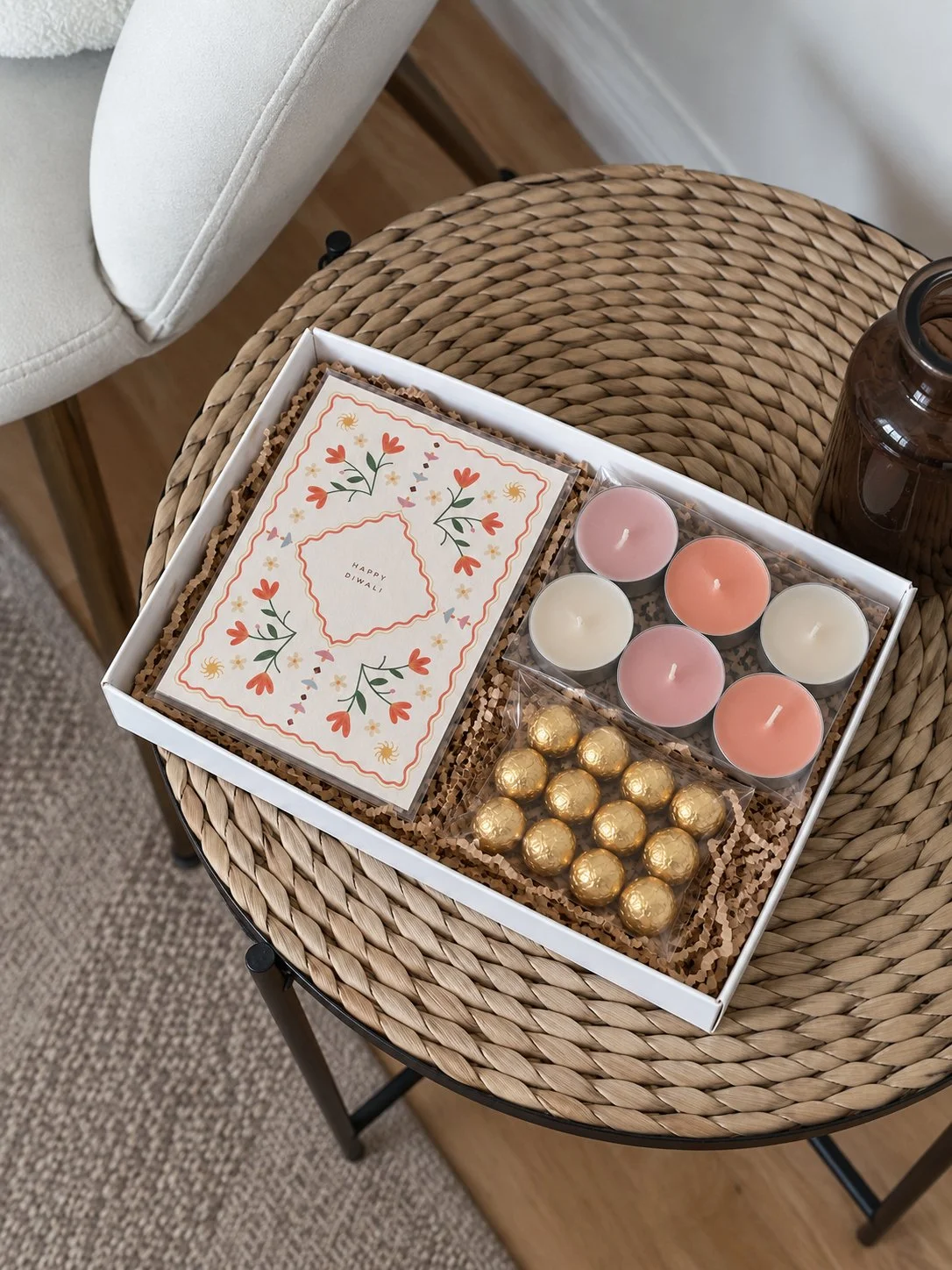 A gift box on a round, woven rattan table containing a decorative card with 'Happy Diwali' and floral designs, six pastel-colored candles, and gold-wrapped chocolates.