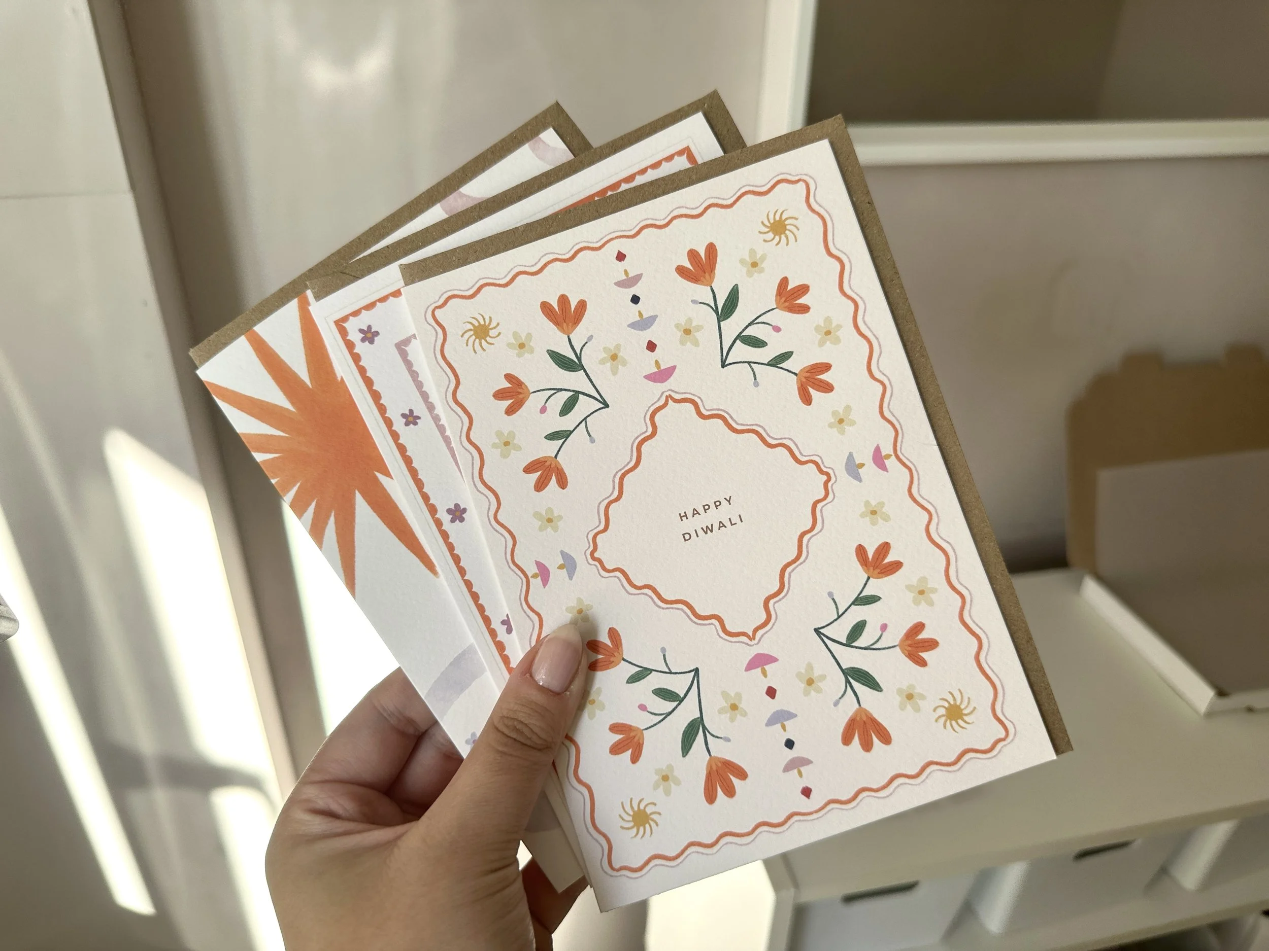 Person holding Diwali greeting cards with floral and sun designs near a window.